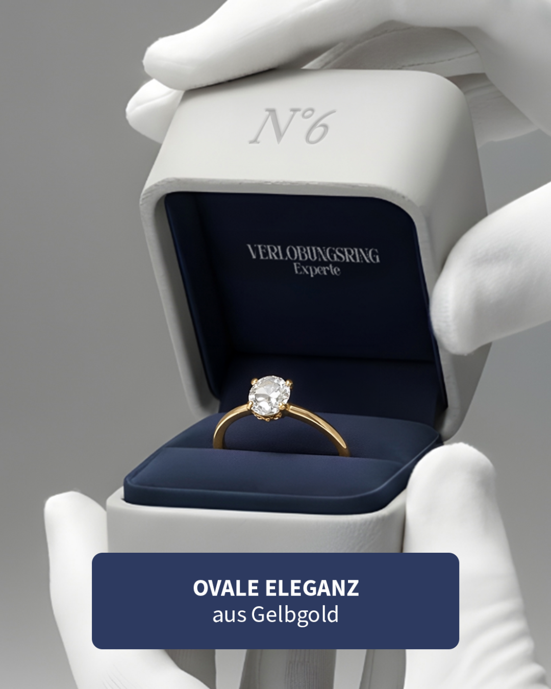 Ein Ring mit ovalem Diamanten in einer blauen Schmuckschachtel, gehalten von weißen Handschuhen. Der Schmuck ist aus Gelbgold, markiert mit 'N°6' auf der Verpackung.