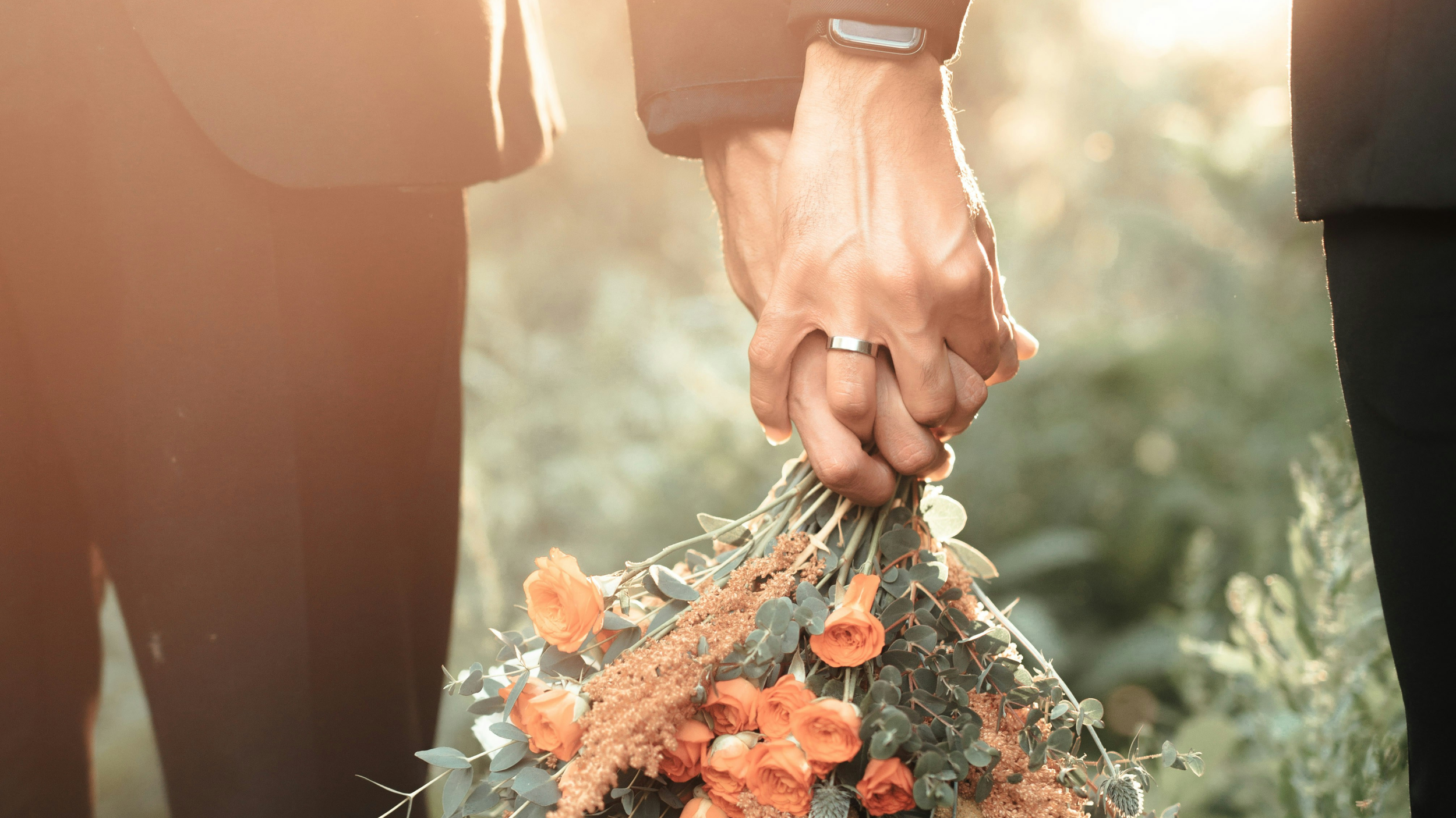 Nahaufnahme eines Paares auf ihrer Hochzeit, das sich an den Händen hält, mit einem Brautstrauß aus orangefarbenen Blumen und Eukalyptus und goldenen Eheringen oder Partnerringen