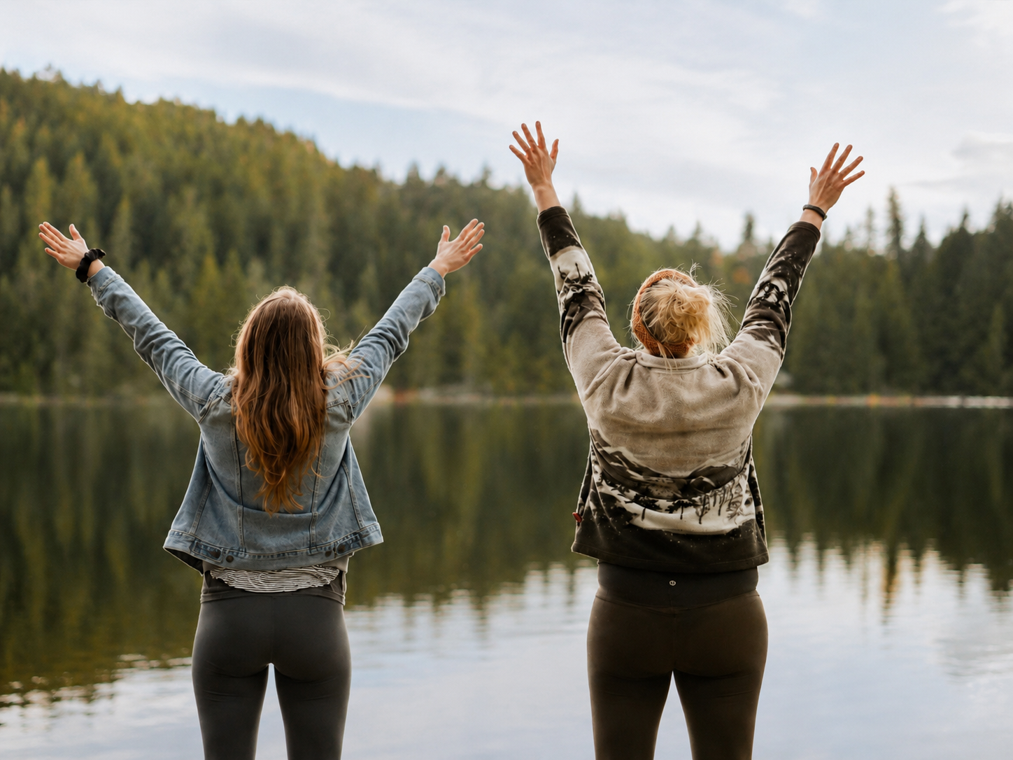 Zwei Frauen stehen am Ufer eines Sees mit erhobenen Armen, Bäume im Hintergrund.