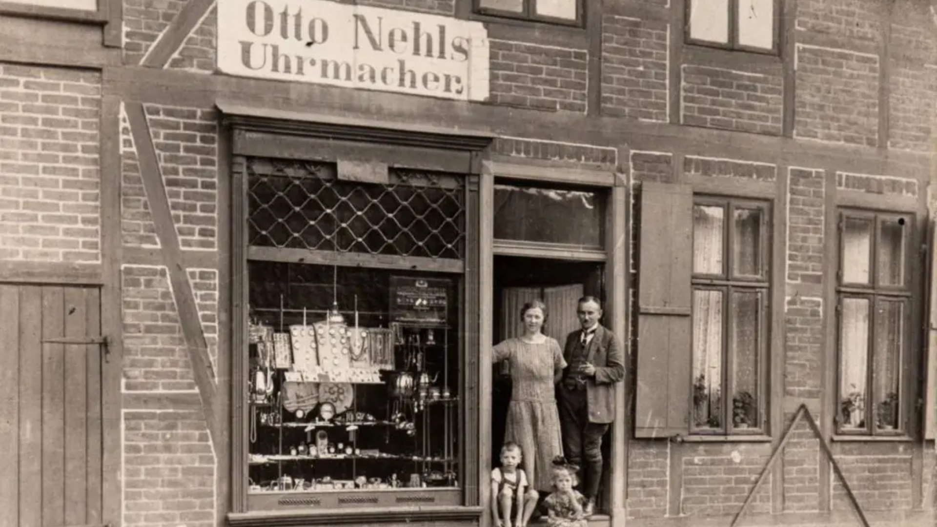 Schwarz-Weiß-Foto einer alten Ladenfront mit einem Schild 'Otto Nehls Uhrmacher'. Vor dem Laden stehen ein Mann, eine Frau und zwei Kinder, die vermutlich eine Familie sind. Im Schaufenster sind Uhren und andere kleine Gegenstände ausgestellt. Das Gebäude besteht aus Ziegelmauerwerk und hat mehrere Fenster.