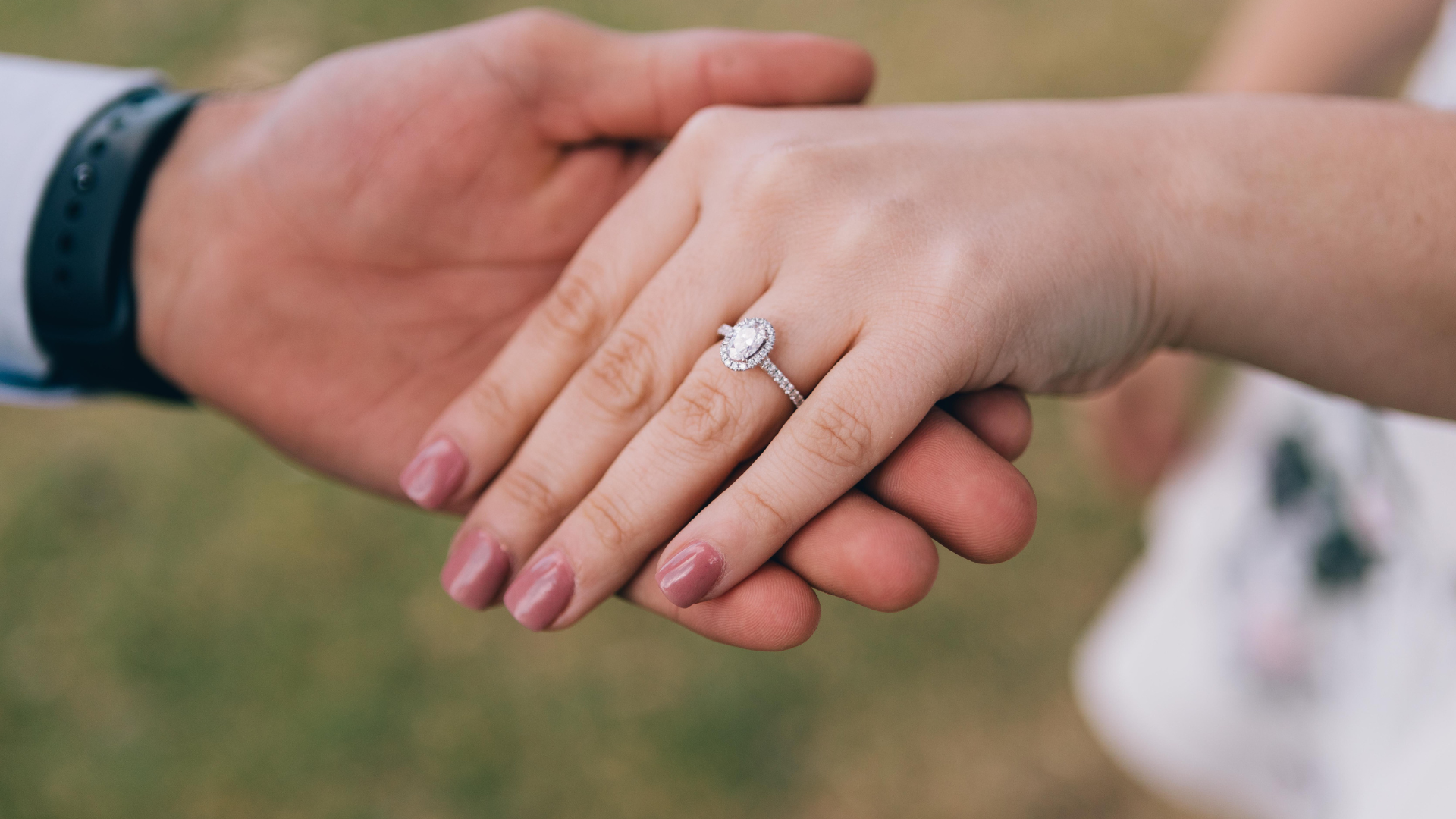 Ein Nahaufnahme-Foto von einem Verlobungsring mit Diamant auf dem Finger einer Frau, die ihre Hand hält, während sie mit ihrem Partner Händchen hält.