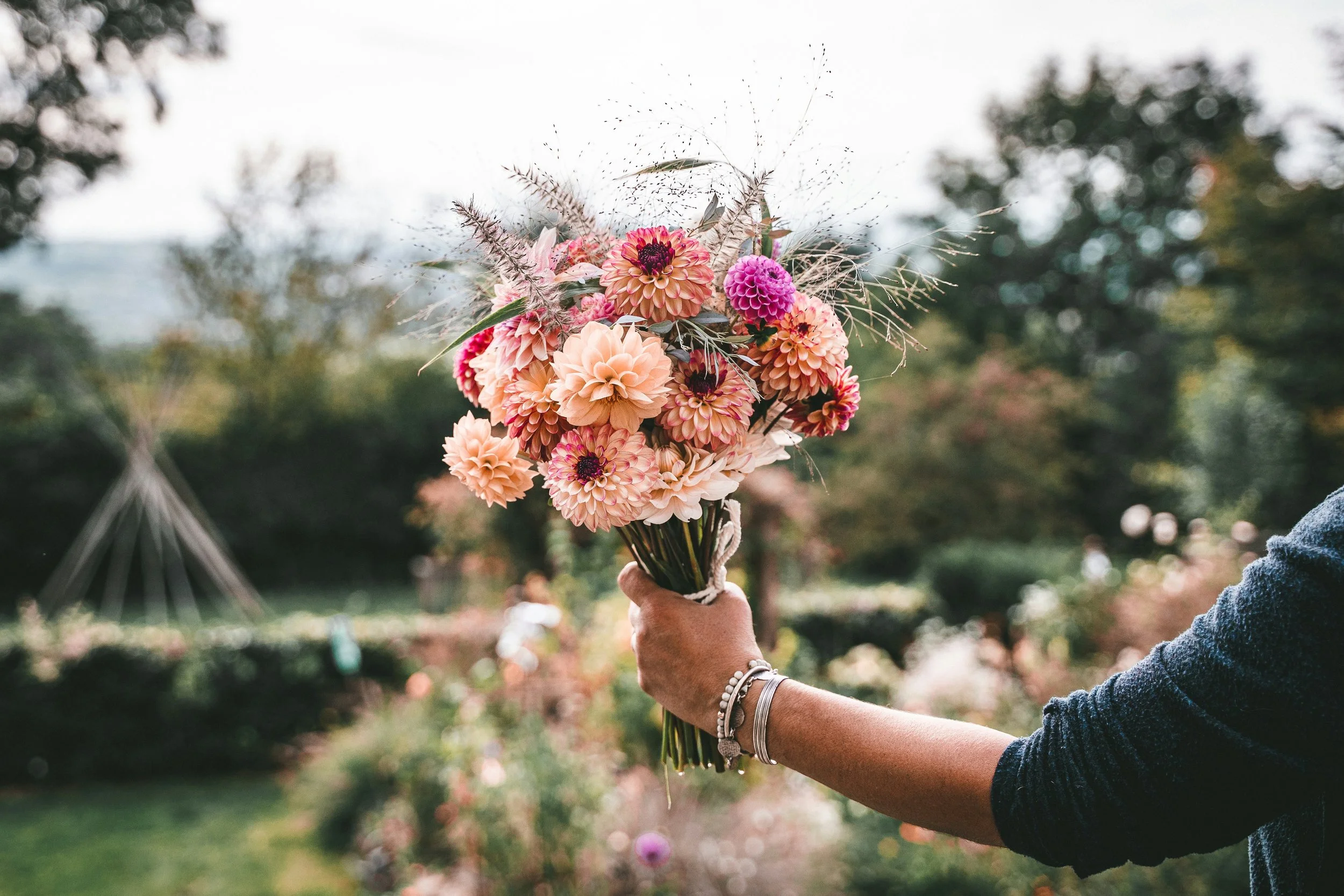 Eine Person hält einen bunten Blumenstrauß im Freien, im Hintergrund sind Bäume und ein Himmel zu sehen.
