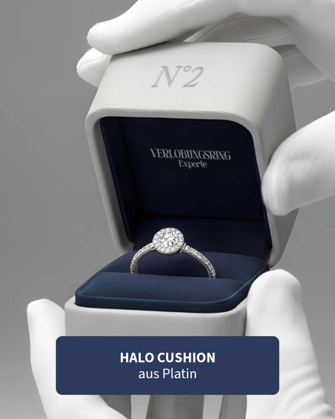 Ringe in einer Schmuckschachtel, die von weißen Handschuhen gehalten wird, mit einem Etikett, das sagt "HALO CUSHION aus Platin".
