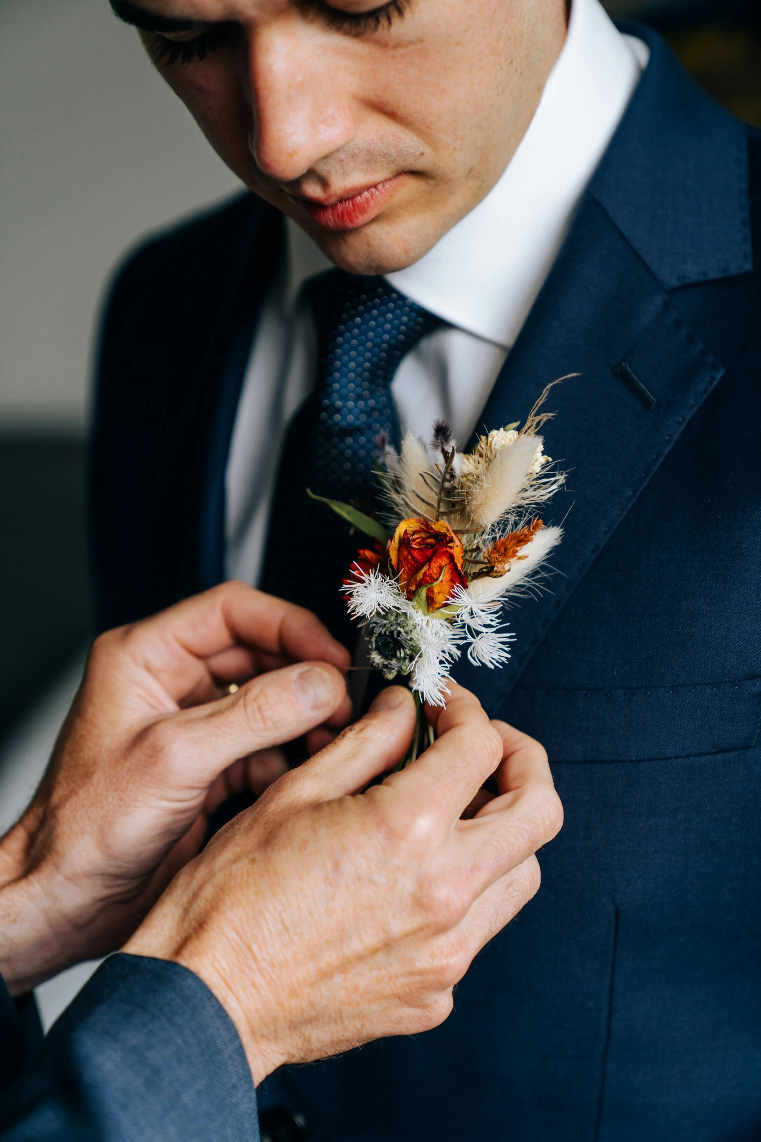 Ein Mann in einem dunklen Anzug und weißem Hemd erhält von einer anderen Person ein Blumenbouquet, vermutlich bei einer formellen Gelegenheit wie einer Hochzeit.