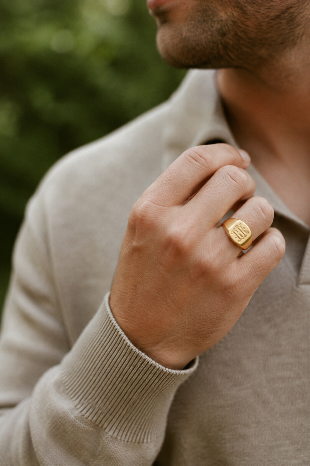 Nahaufnahme einer männlichen Hand, die eine goldene Siegelring trägt, die in die Nähe des Halses einer Person gehalten wird, mit einem grauen Hemd und einem beige Pullover. Der Hintergrund ist unscharf und grün.