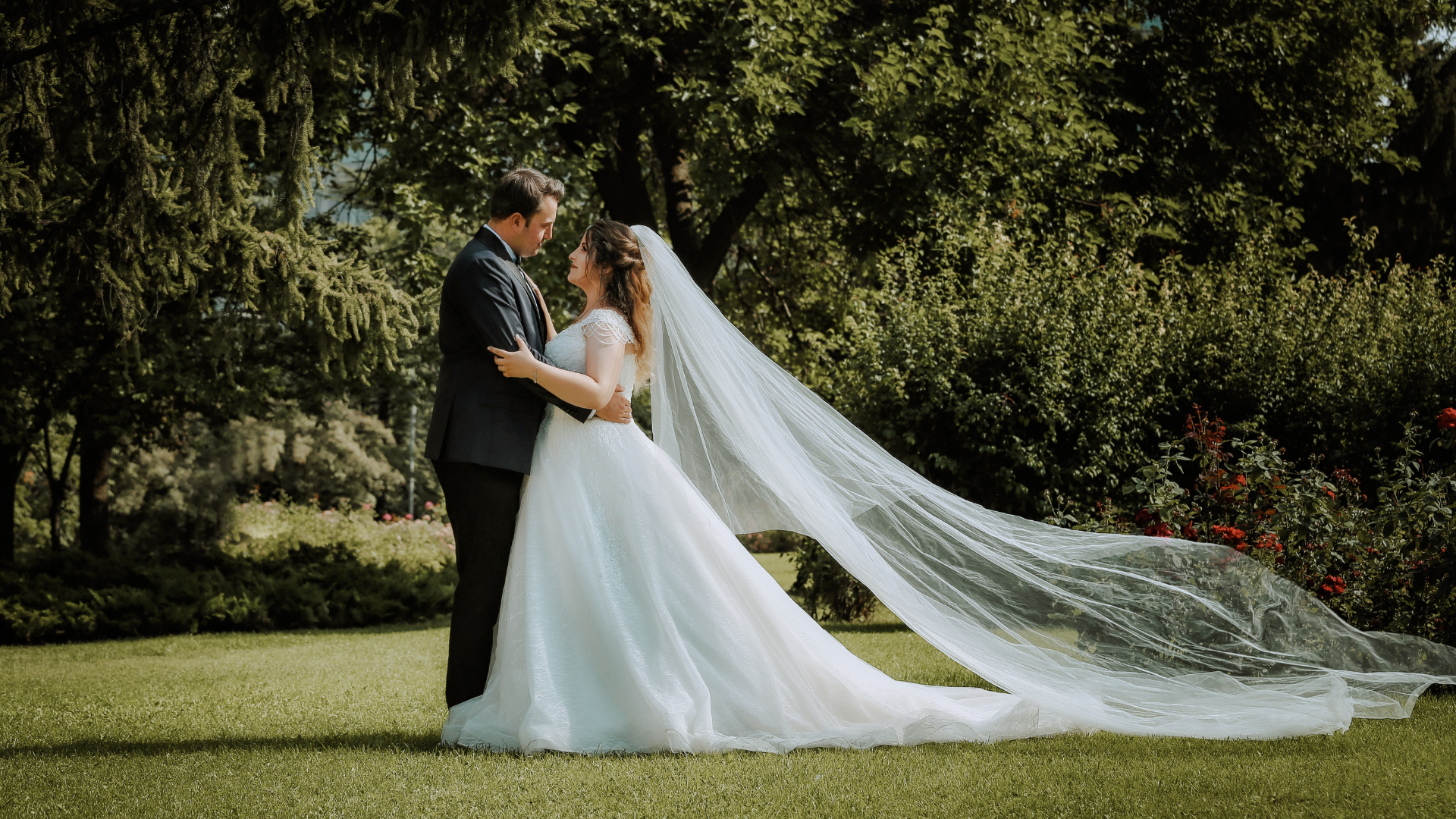 Ein Brautpaar steht sich in an ihrer Hochzeit einem Garten gegenüber, umarmt sich und blickt sich liebevoll an, während das Brautkleid und der Schleier im Wind wehen und ihre Trauringe in der Sonne funkeln.