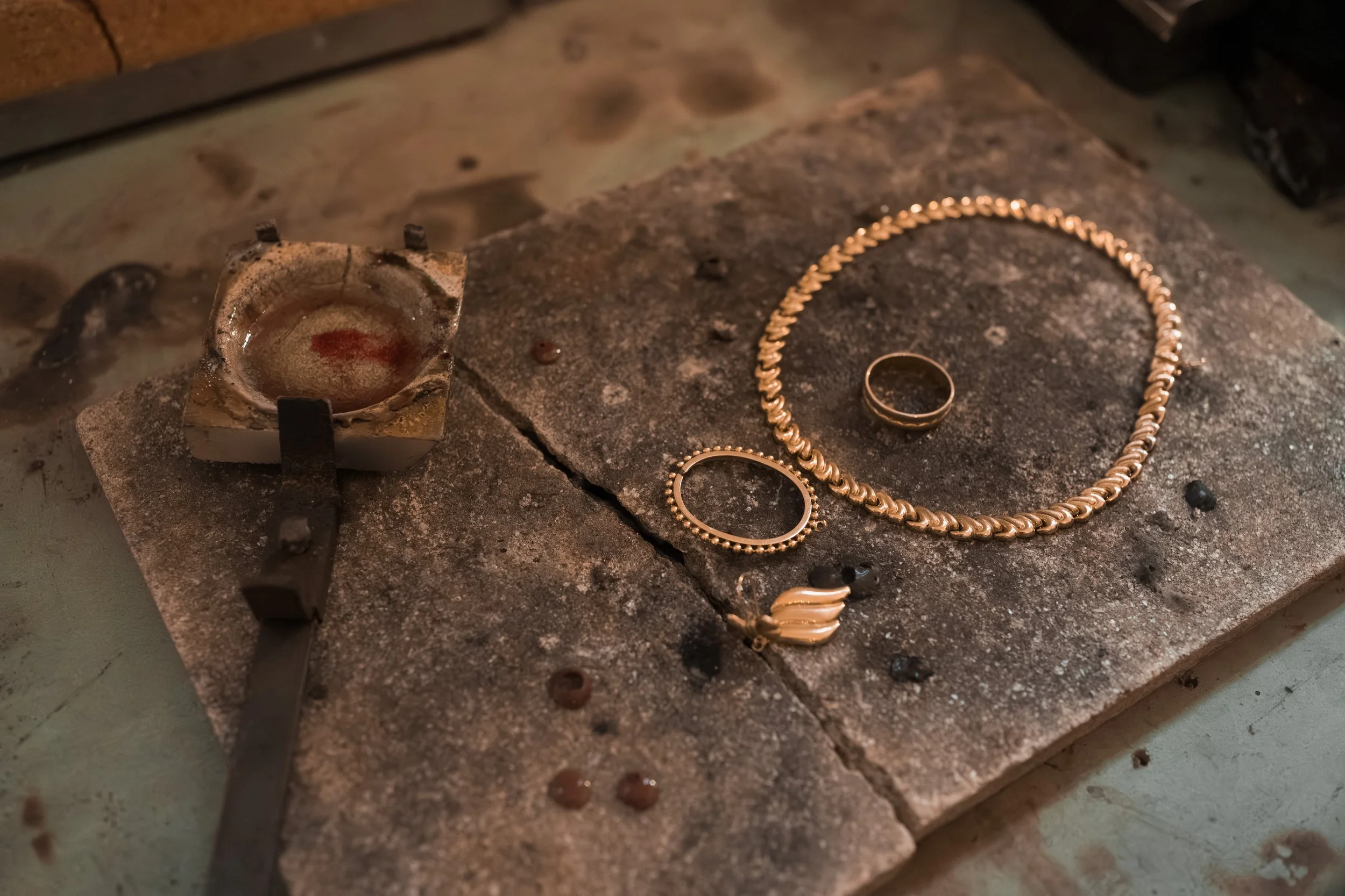 Goldschmiedewerkstatt mit Schmuckstücken, wie einer Kette, Armband und Ring, die auf einer Werkbank liegen. Eine erhitzte Metallform ist ebenfalls sichtbar.