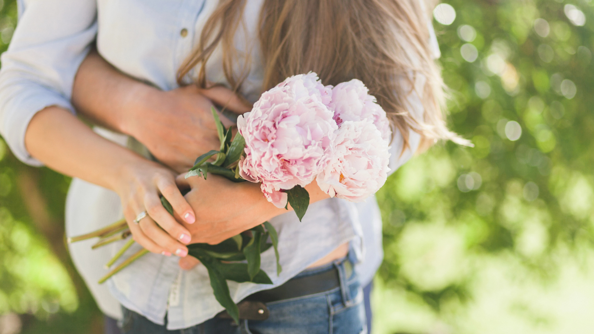 Eine Person trägt einen Ehering und einen Verlobungsring und hält einen Strauß rosa Pfingstrosen im Freien, mit grünen Bäumen im Hintergrund.