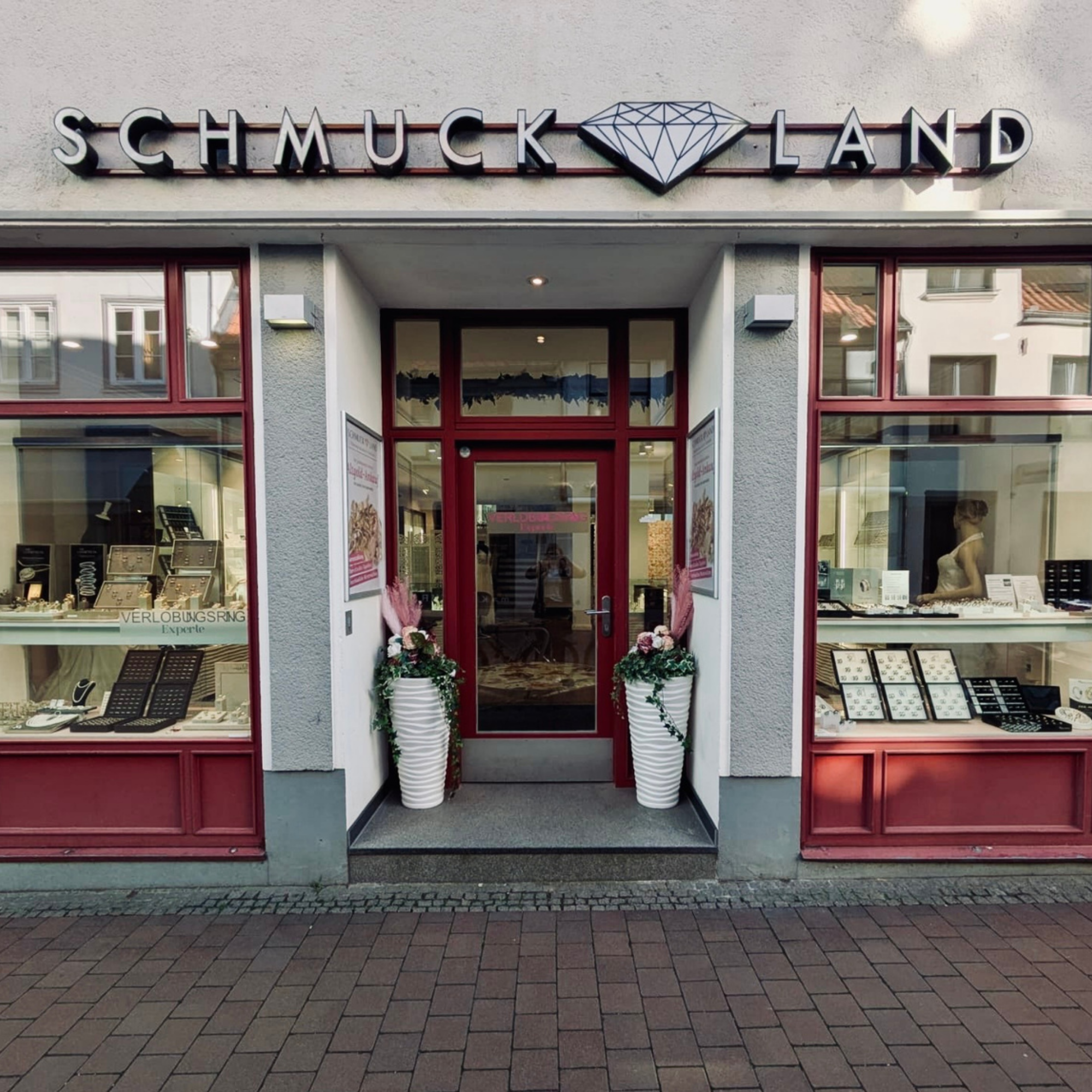 Schmuckladen mit Schmuckdisplay im Schaufenster, zwei große weiße Blumenkübel mit Blumen vor der Tür, rote Türrahmen, Fenster mit Schmuckstücken. Außenaufnahme