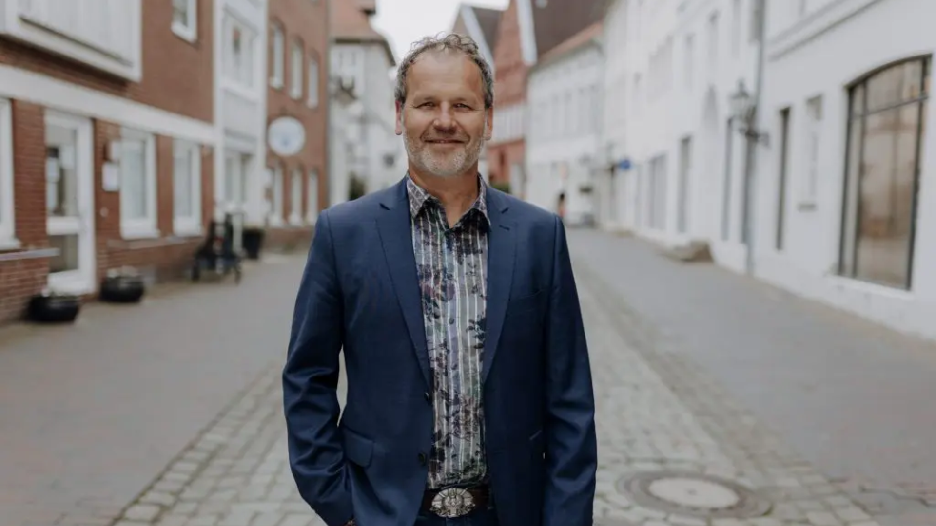 Ein Mann in einem blauen Anzug und kariertem Hemd steht auf einer gepflasterten Straße vor einem historischen Stadthaus mit weißen und roten Backsteinfassaden.
