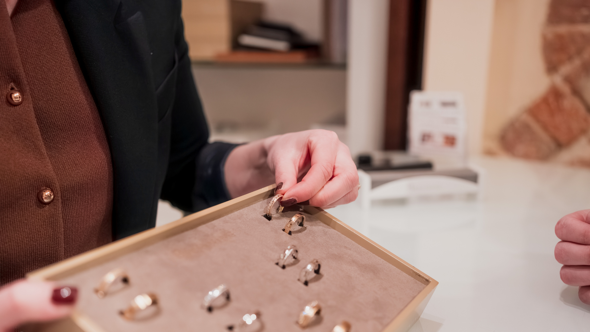 Nahaufnahme einer Person, die Ringe in einem Schmuckladen betrachtet. Die Person hält einen Ring in der Hand, während andere Ringe in einer Schmuckschatulle auf einem Tisch liegen.