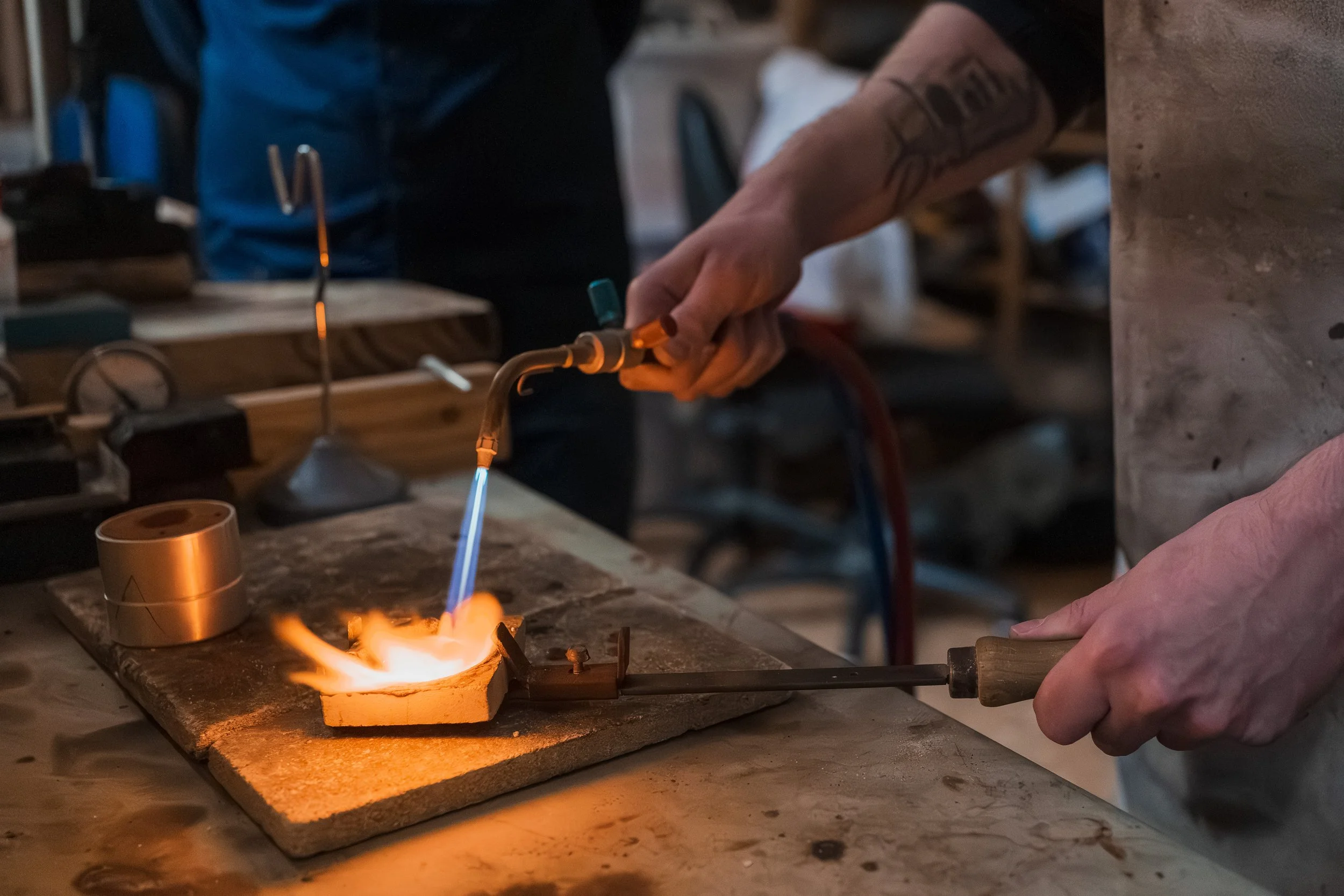 Ein Mensch arbeitet in einer Werkstatt mit einem Lötgerät an einer Metallarbeit. Die Flamme ist sichtbar, die Person trägt eine Tätowierung am Arm und trägt Arbeitshandschuhe.