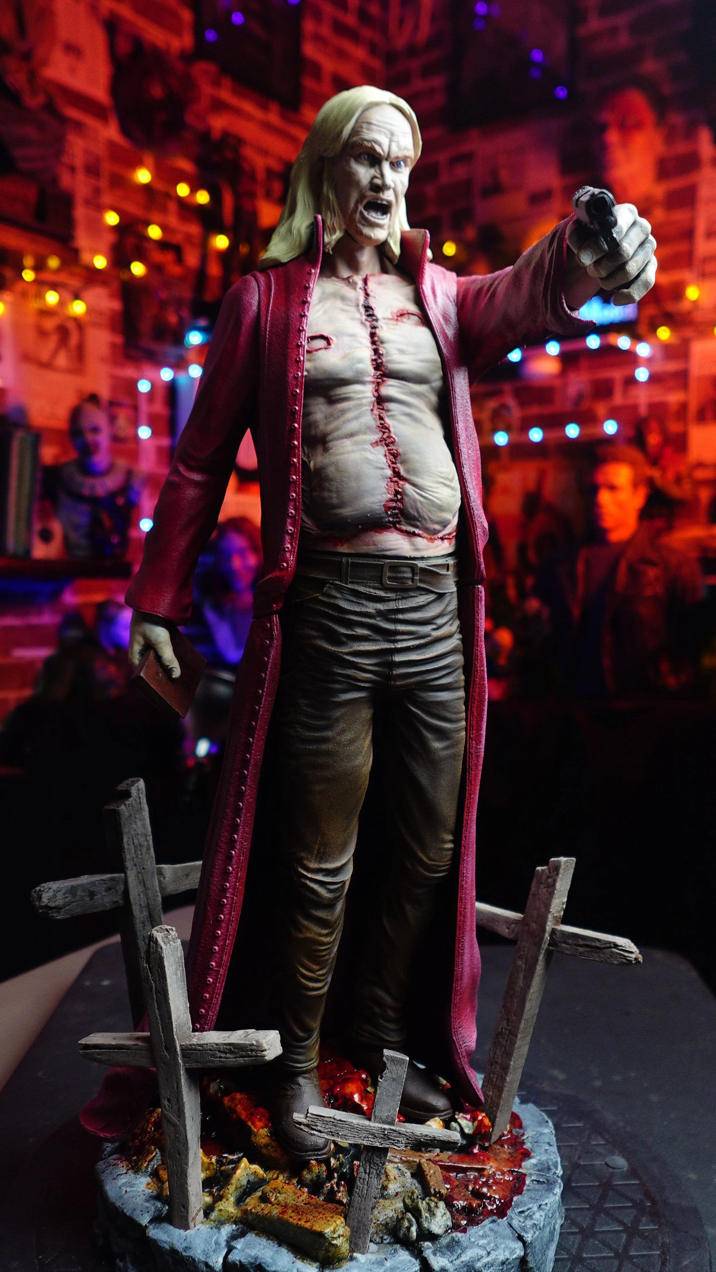 A detailed sculpture of Negan, a character from The Walking Dead, holding a gun and standing amidst broken wooden carts and debris on fire, with a lively background of string lights and posters.
