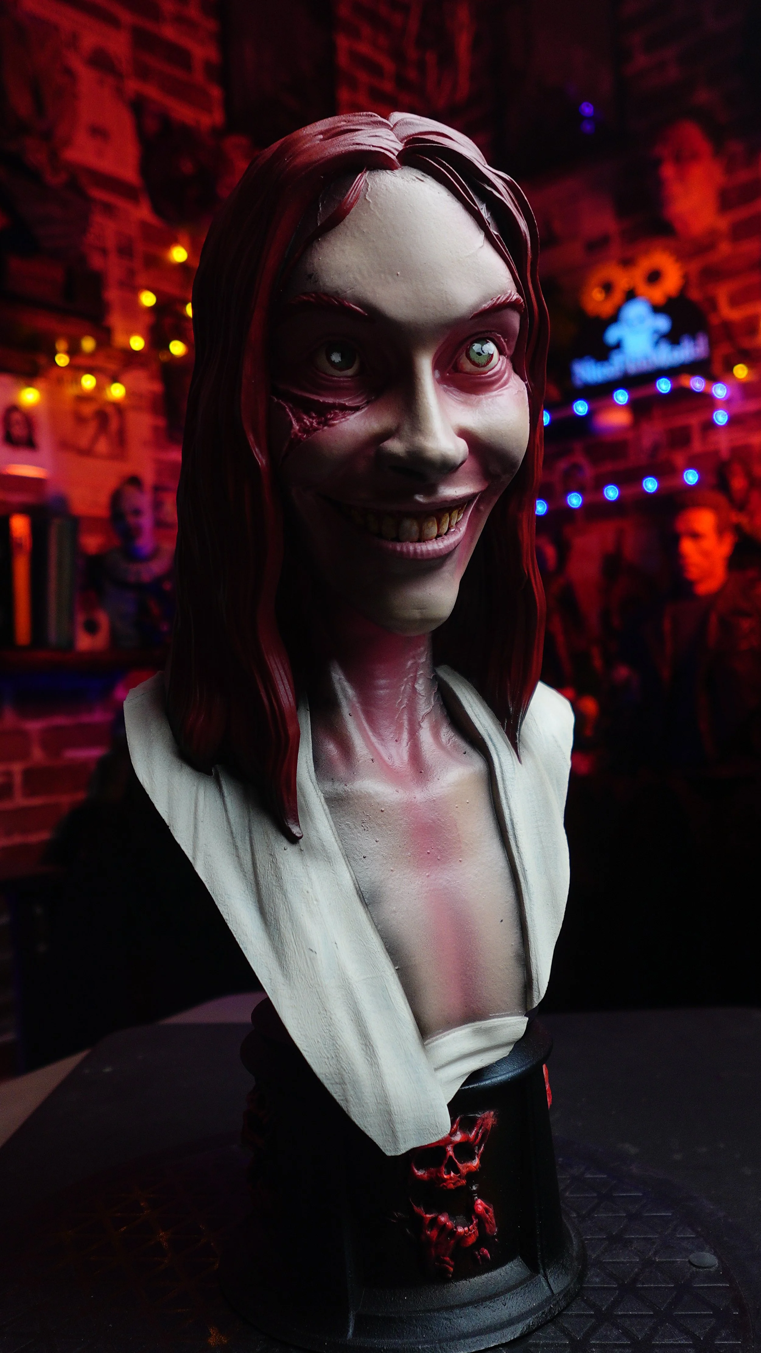 A Halloween-themed bust of a woman with devil-like features, including pale skin, long red hair, glowing eyes, and a sinister smile, set on a black platform with red skull decorations, in a dimly lit room with red and blue neon lights and Halloween d