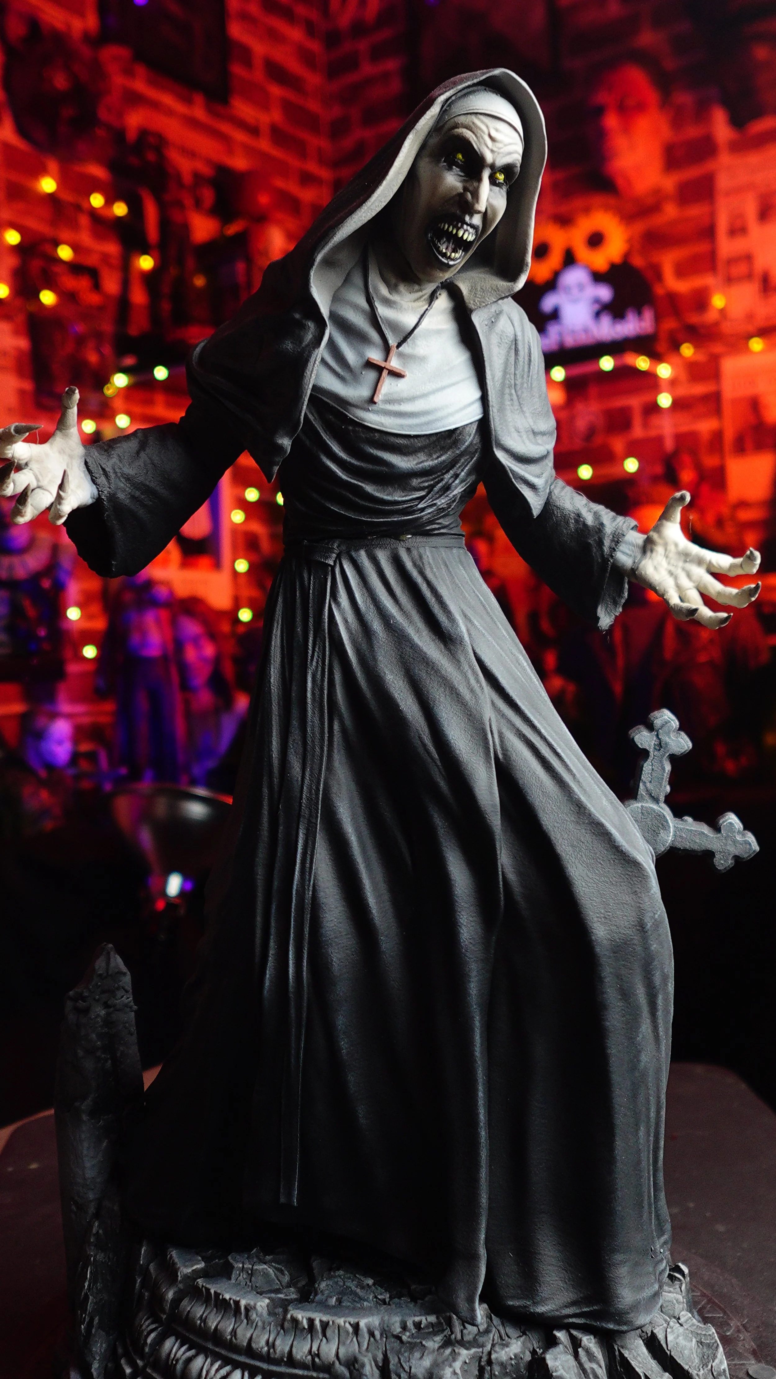 A sinister-looking gothic sculpture of a nun with yellow eyes, bared teeth, and a menacing expression, dressed in a traditional nun's habit, with a graveyard setting including a tombstone and cross in the foreground, illuminated by colorful Halloween
