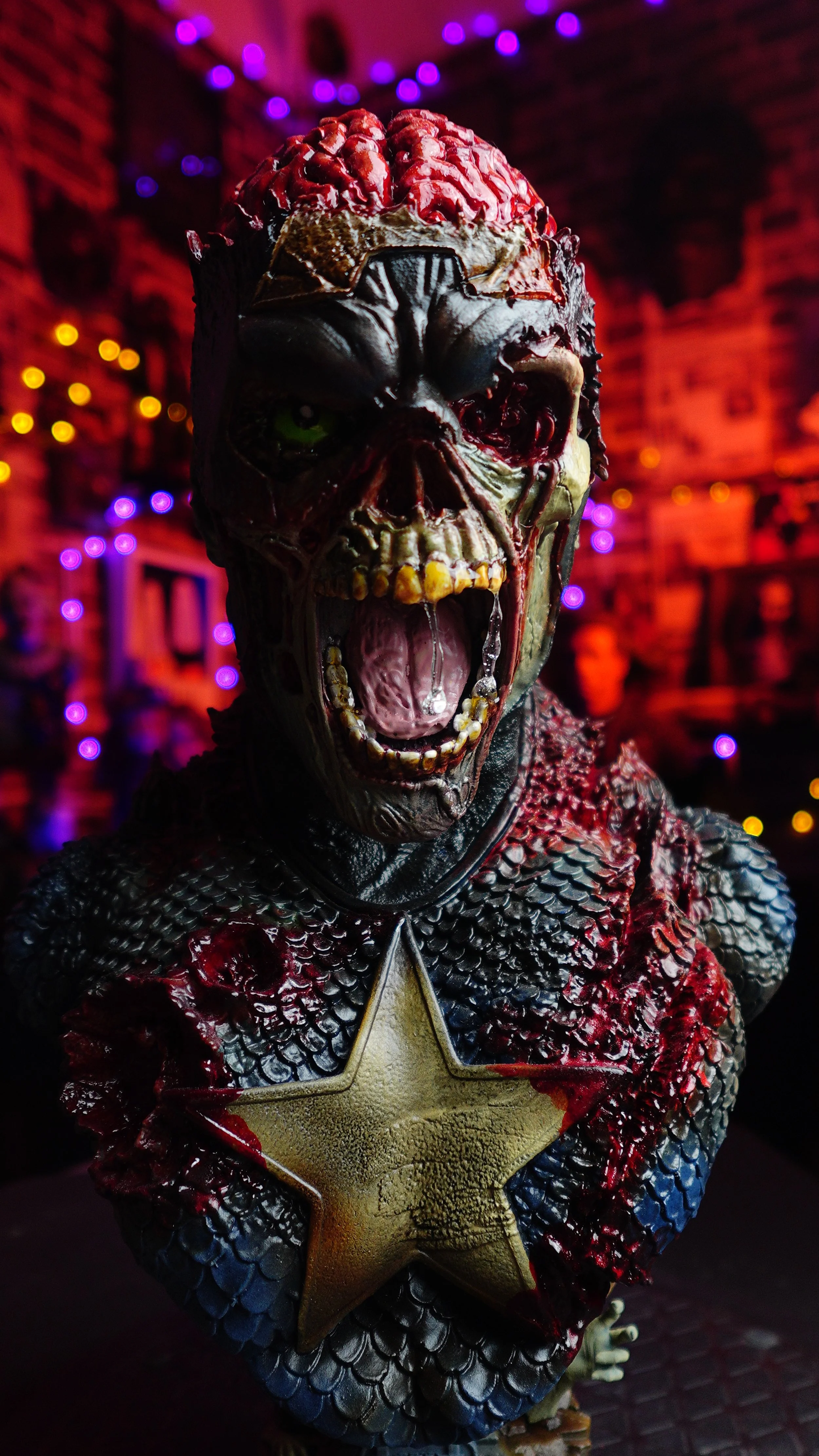A detailed zombie-themed sculpture of a creature with a partially exposed brain, sharp teeth, and half of its face decayed. The creature is wearing a costume resembling Captain America's shield on its chest, set against a colorful background with pur