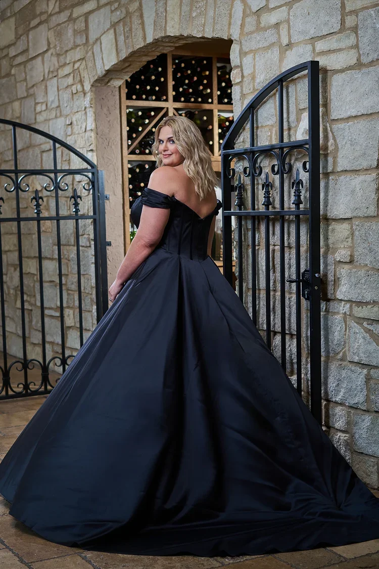 Woman in a black off-shoulder evening gown standing by an open wrought iron gate, with a stone wall and a wine cellar in the background.