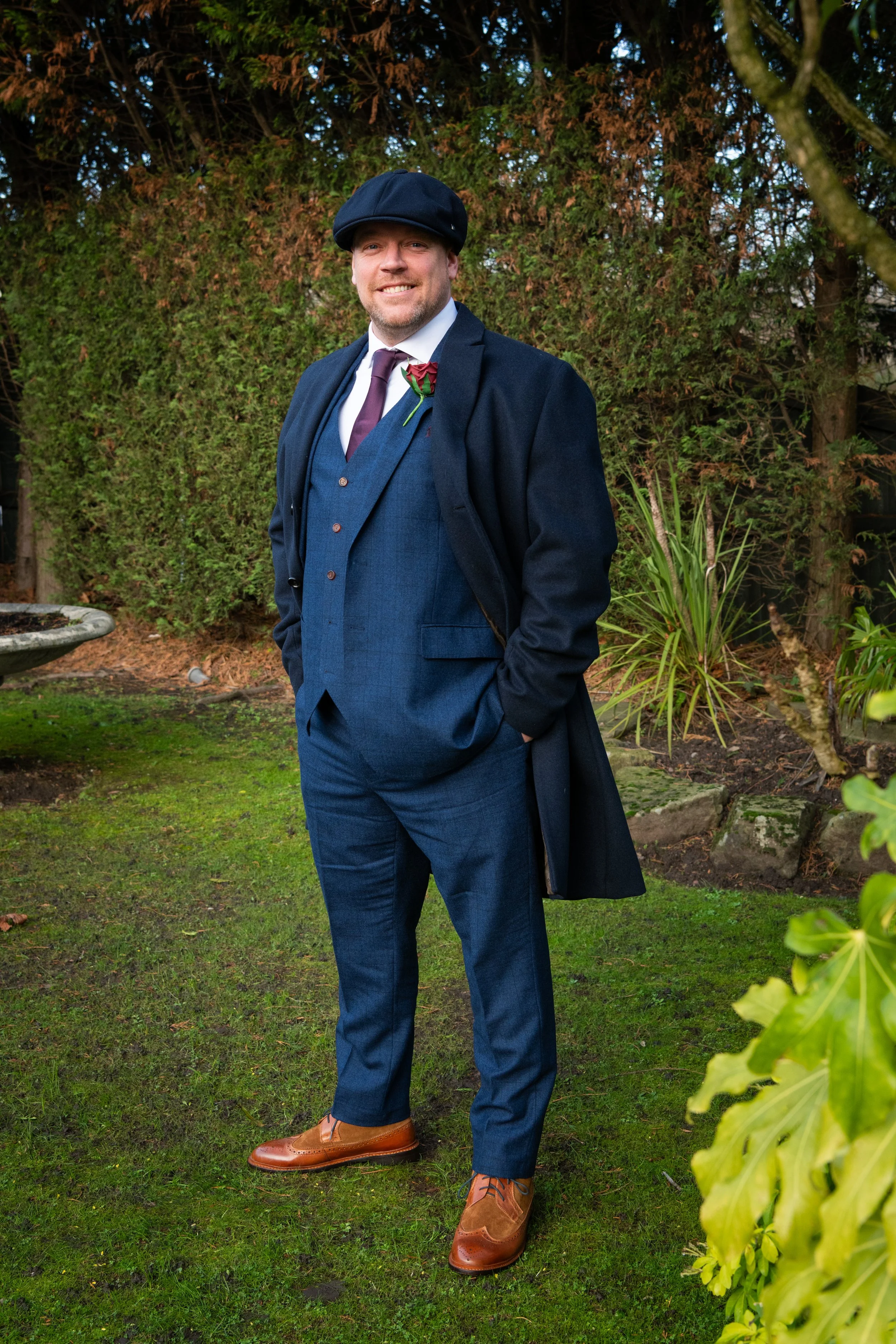 A man in a dark blue suit, brown shoes, purple tie, and dark overcoat standing outdoors on a grassy area with trees and bushes in the background. He is smiling and wearing a flat cap, with his hands in his pockets.