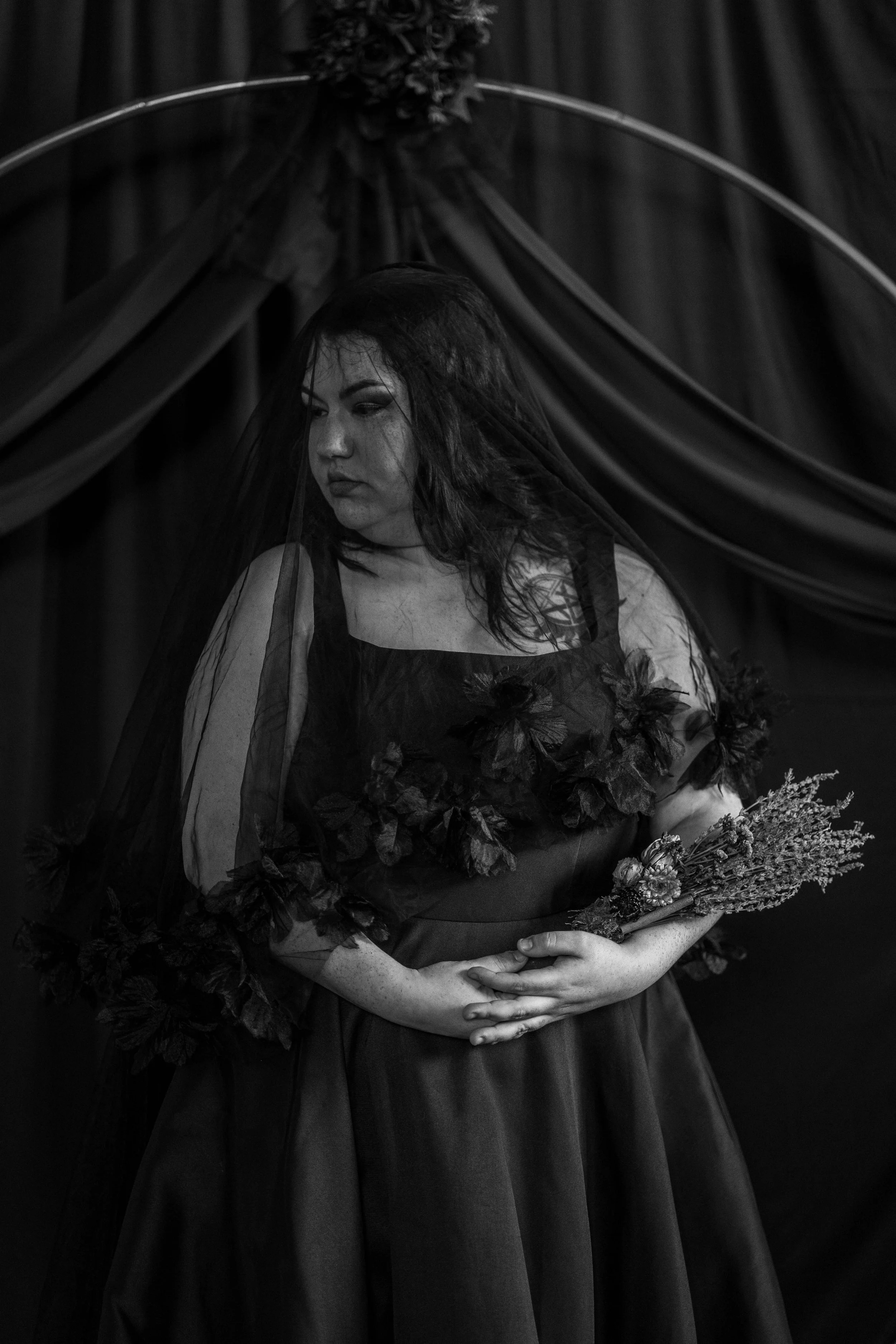A woman dressed in dark attire with a veil, holding a bouquet of flowers, standing in front of a curtain with draped fabric and floral decorations, in black and white photography.