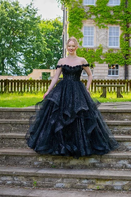 A young woman in a black, off-the-shoulder ball gown standing on outdoor stone steps with a grassy area, trees, a wooden fence, and a building covered in green ivy in the background.