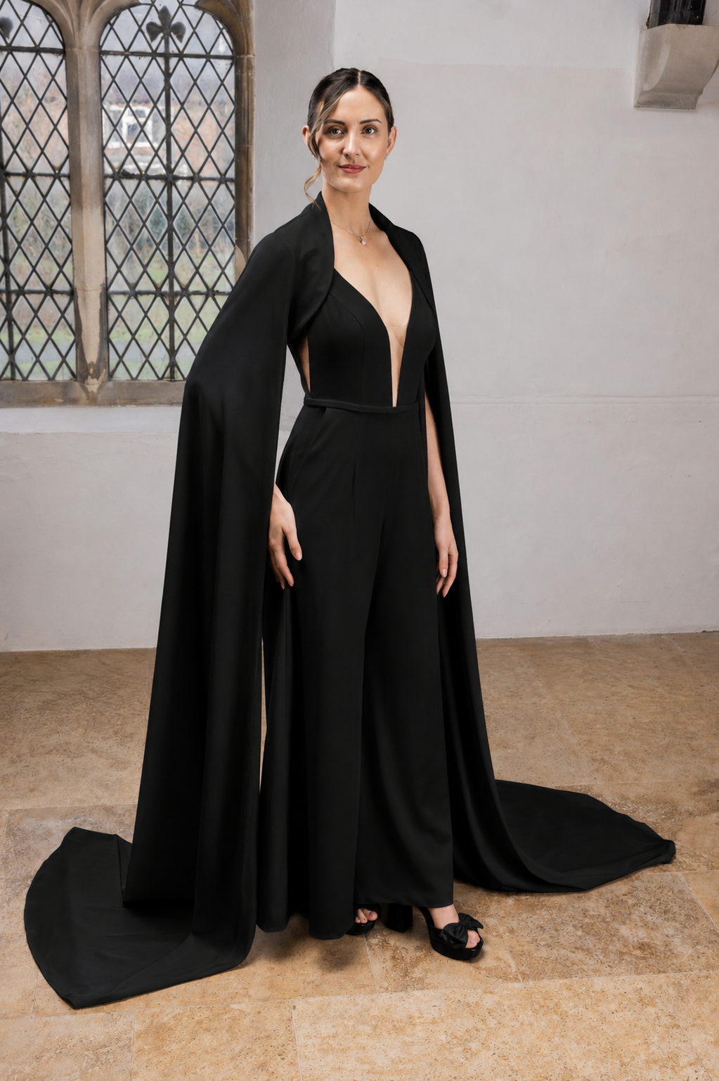 A woman dressed in a black deep V-neck jumpsuit with wide-legged pants, accessorized with a black cape, standing indoors near a window with leaded glass panes.