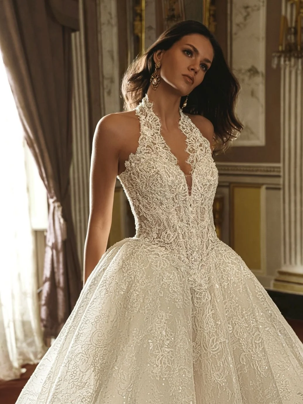 Woman wearing a detailed white lace wedding gown with a full skirt, standing in a richly decorated room with gold accents and draped curtains.