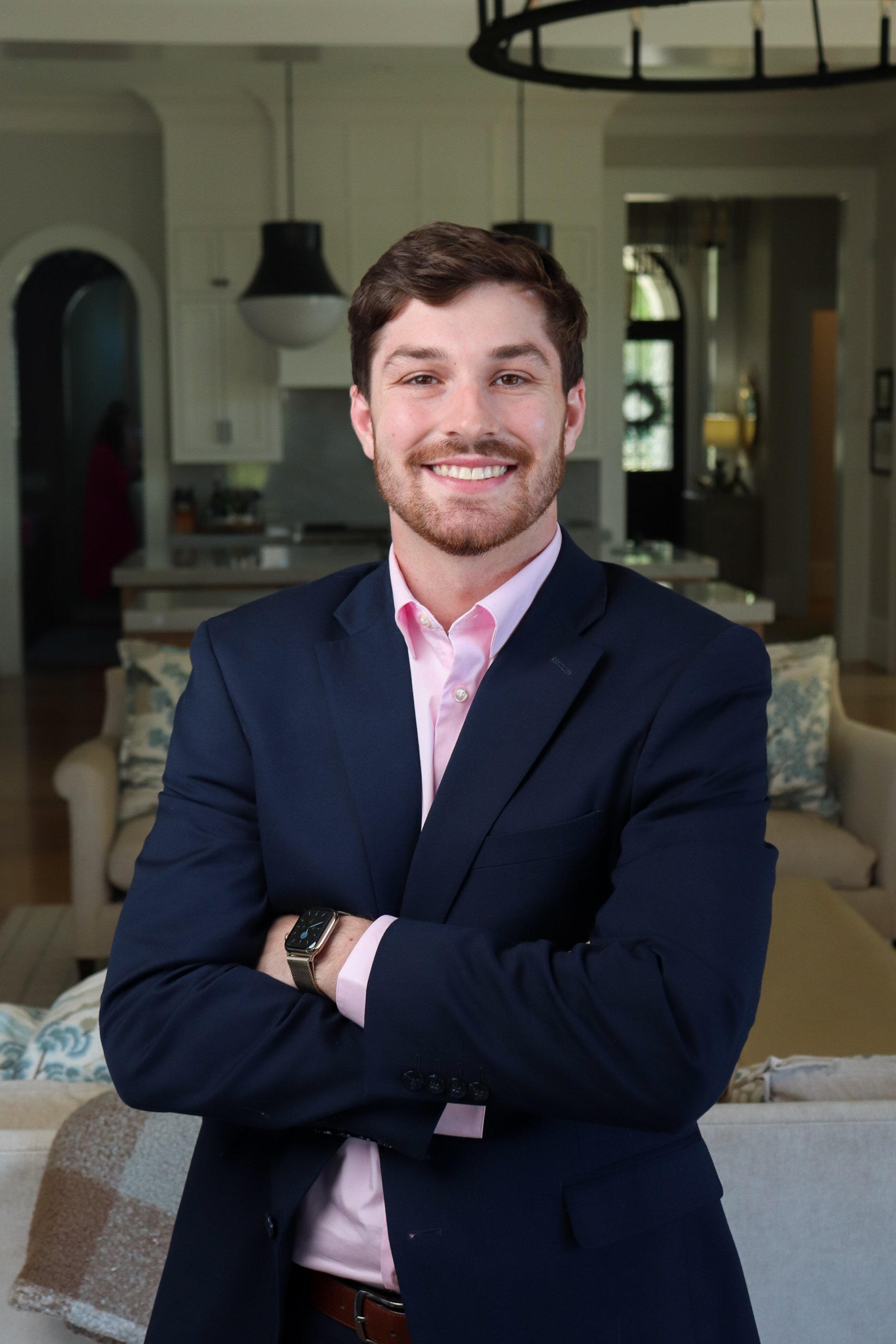 A young man with brown hair, light skin, and a beard is smiling and standing with arms crossed in a living room. He is wearing a dark blue suit, pink shirt, and a watch. The background shows a kitchen and a sofa with cushions.