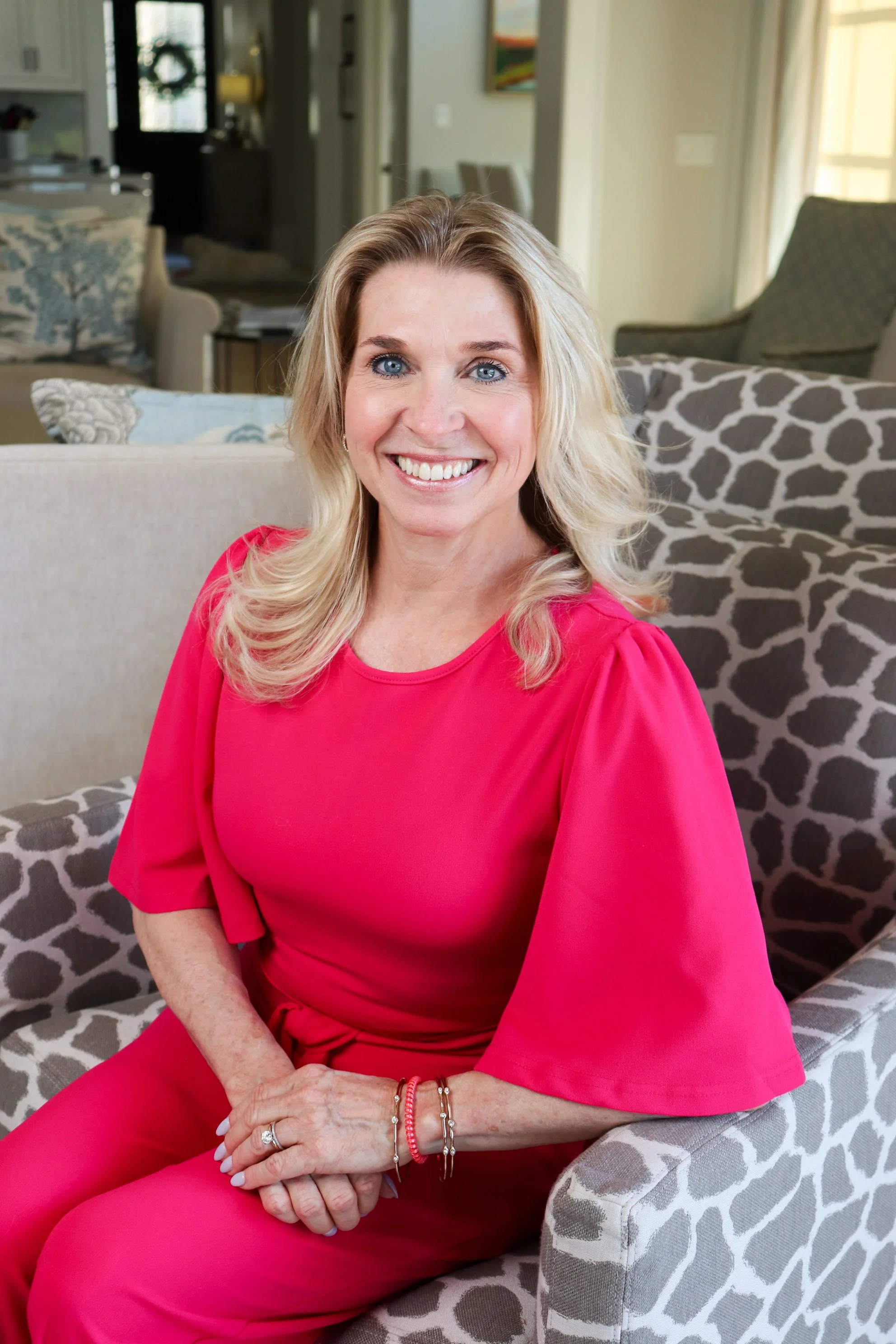 A woman with long blonde hair, blue eyes, and a bright smile sitting on a patterned armchair in a well-lit living room.