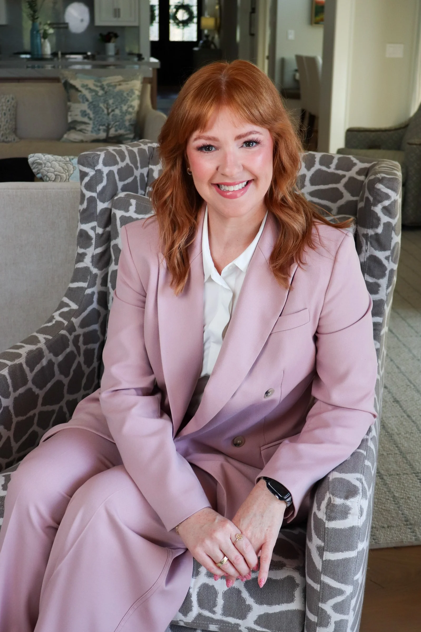 A woman with red hair, wearing a light pink suit, sitting on a patterned armchair with a cheerful expression.