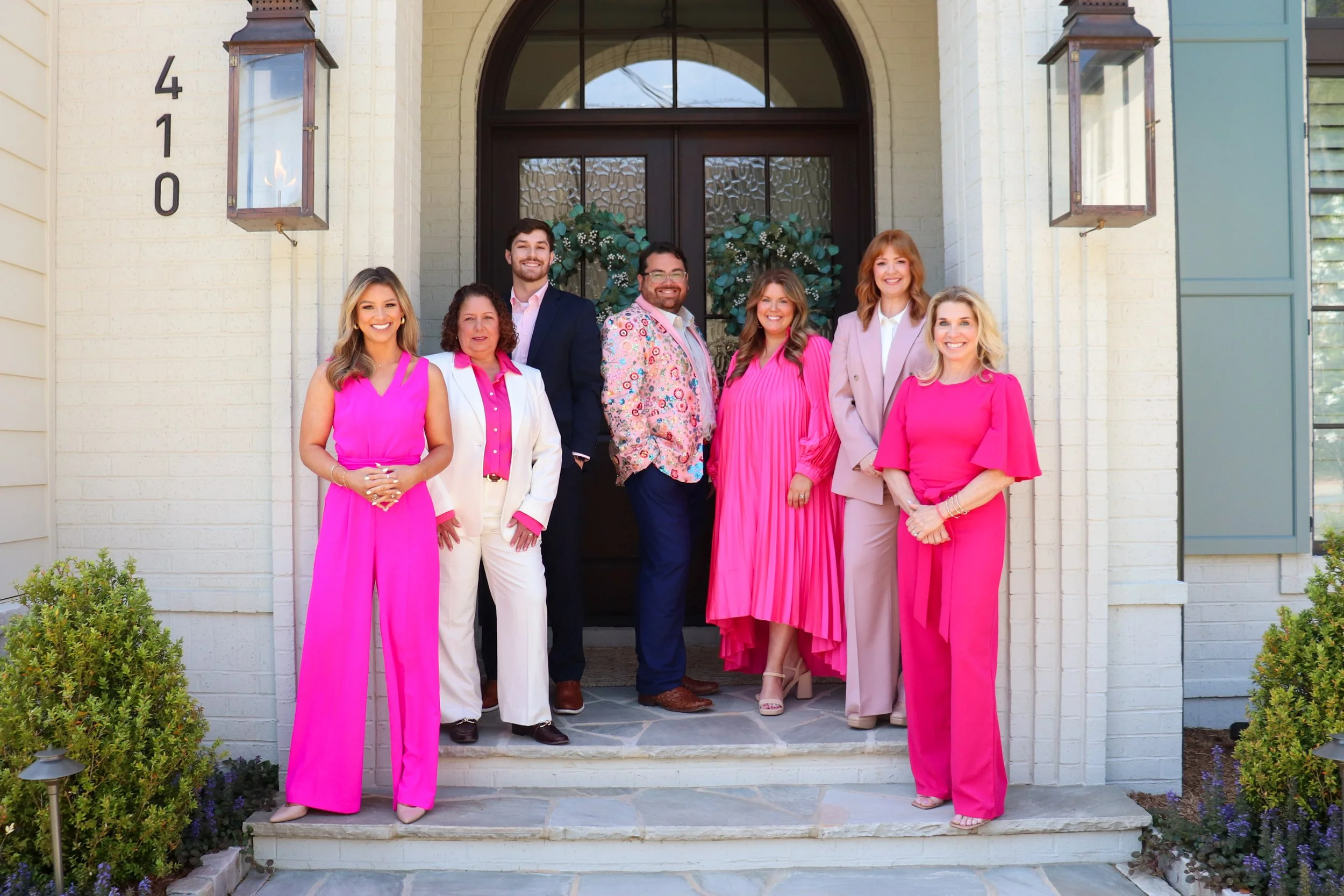 Group of seven people standing on the front steps of a house, smiling for a photo. They are dressed in colorful, mostly pink and pastel outfits, with three women wearing pink dresses and four men in various suits and shirts. The house has a dark front door, a flower wreath hanging on it, and the house number 410 displayed on the wall to the left. There are bushes and small plants around the porch.