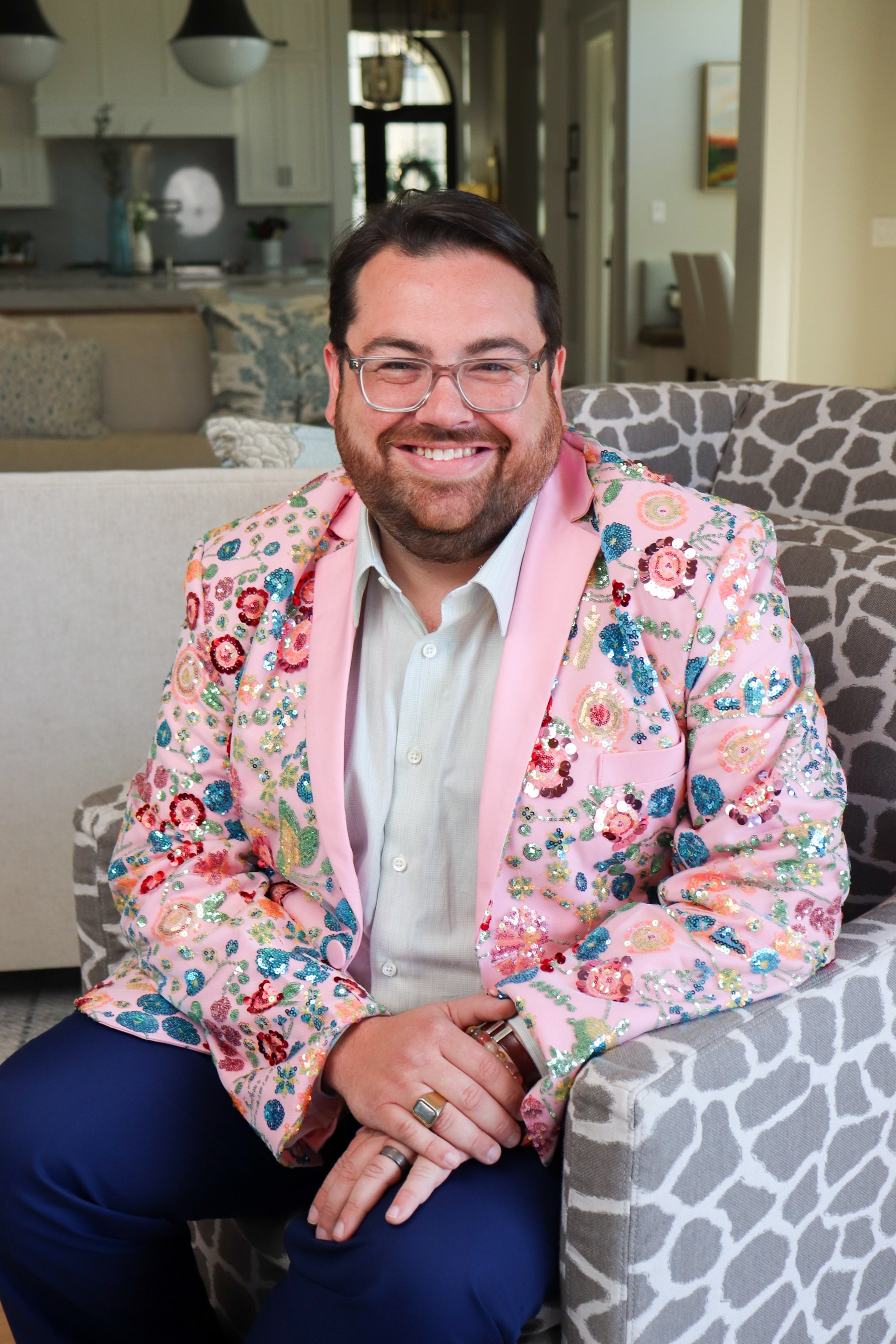 A man with dark hair, glasses, and a beard smiling, wearing a pink blazer adorned with colorful sequined floral patterns, sitting on a patterned chair in a living room with a neutral sofa, decorative pillows, and modern decor.