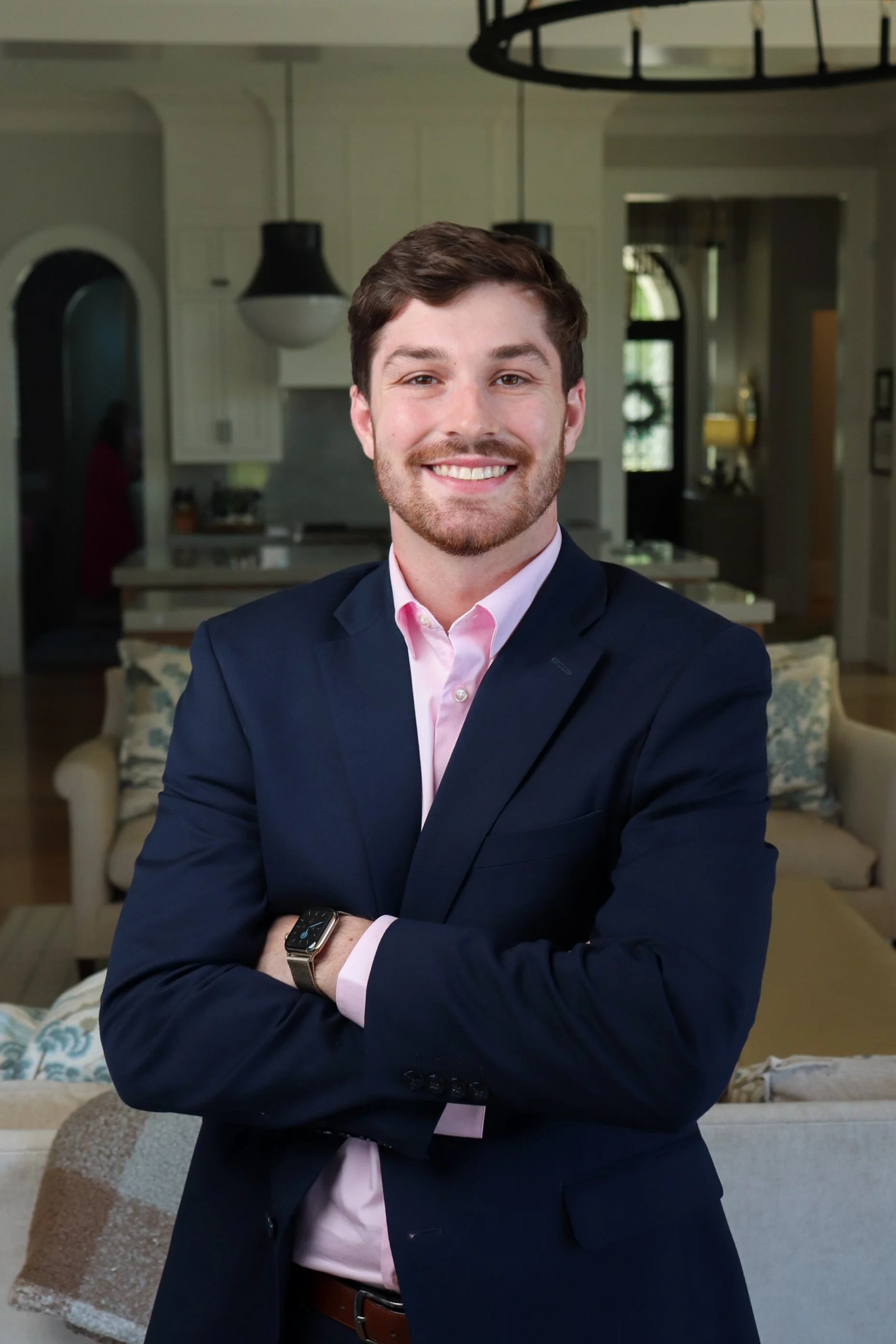 A young man with brown hair and a beard, wearing a navy suit and a light pink shirt, standing with arms crossed and smiling in a bright, modern living room.