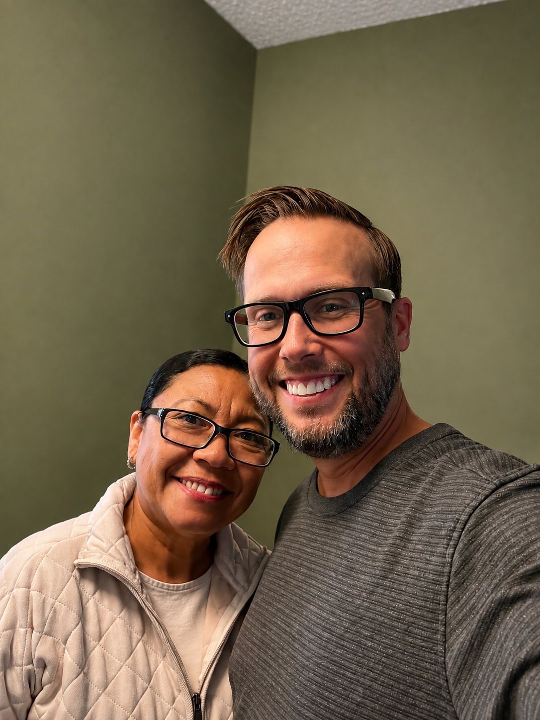 A smiling woman with glasses and a quilted beige jacket standing next to a smiling man with beard, glasses, and gray shirt, both taking a selfie indoors against a green wall.