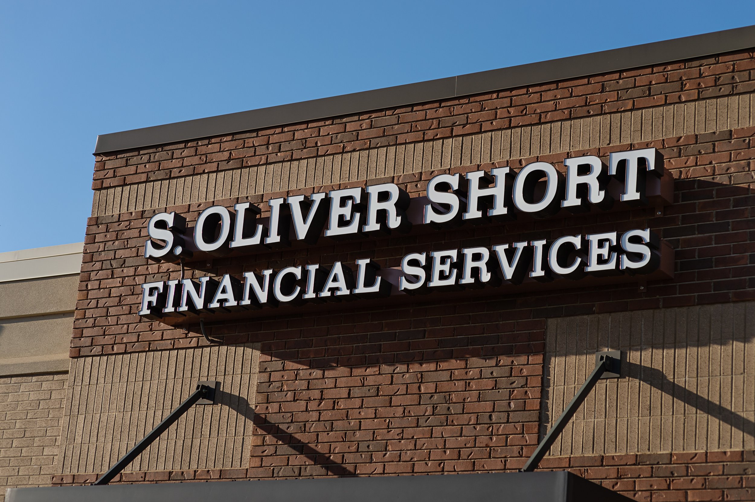 Close-up of a brick commercial building facade with white three-dimensional sign saying "S. Oliver Short Financial Services" against blue sky.