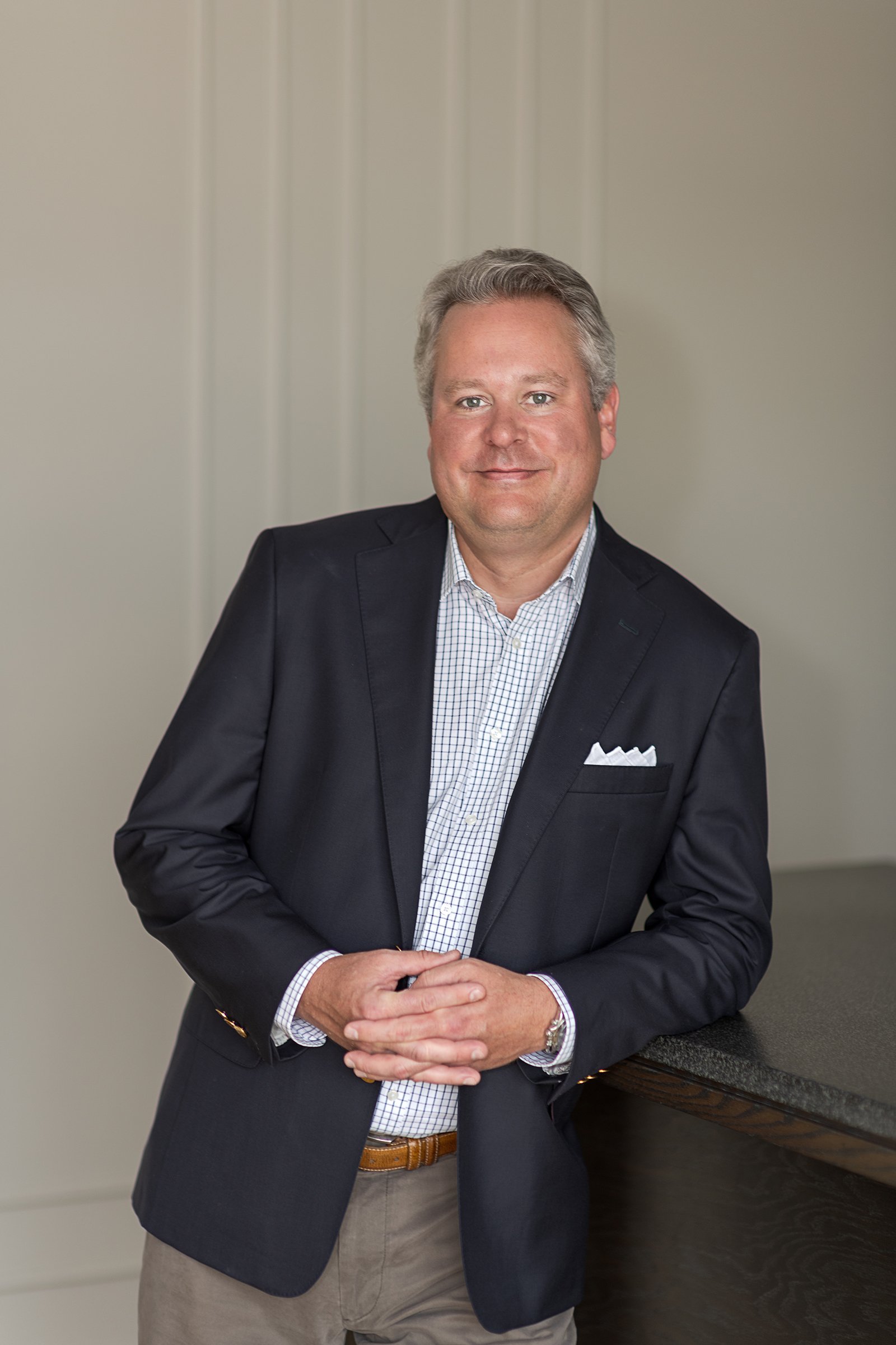 A man with gray hair wearing a dark blazer, a checked shirt, and beige pants, standing indoors with a neutral background and a dark countertop nearby.