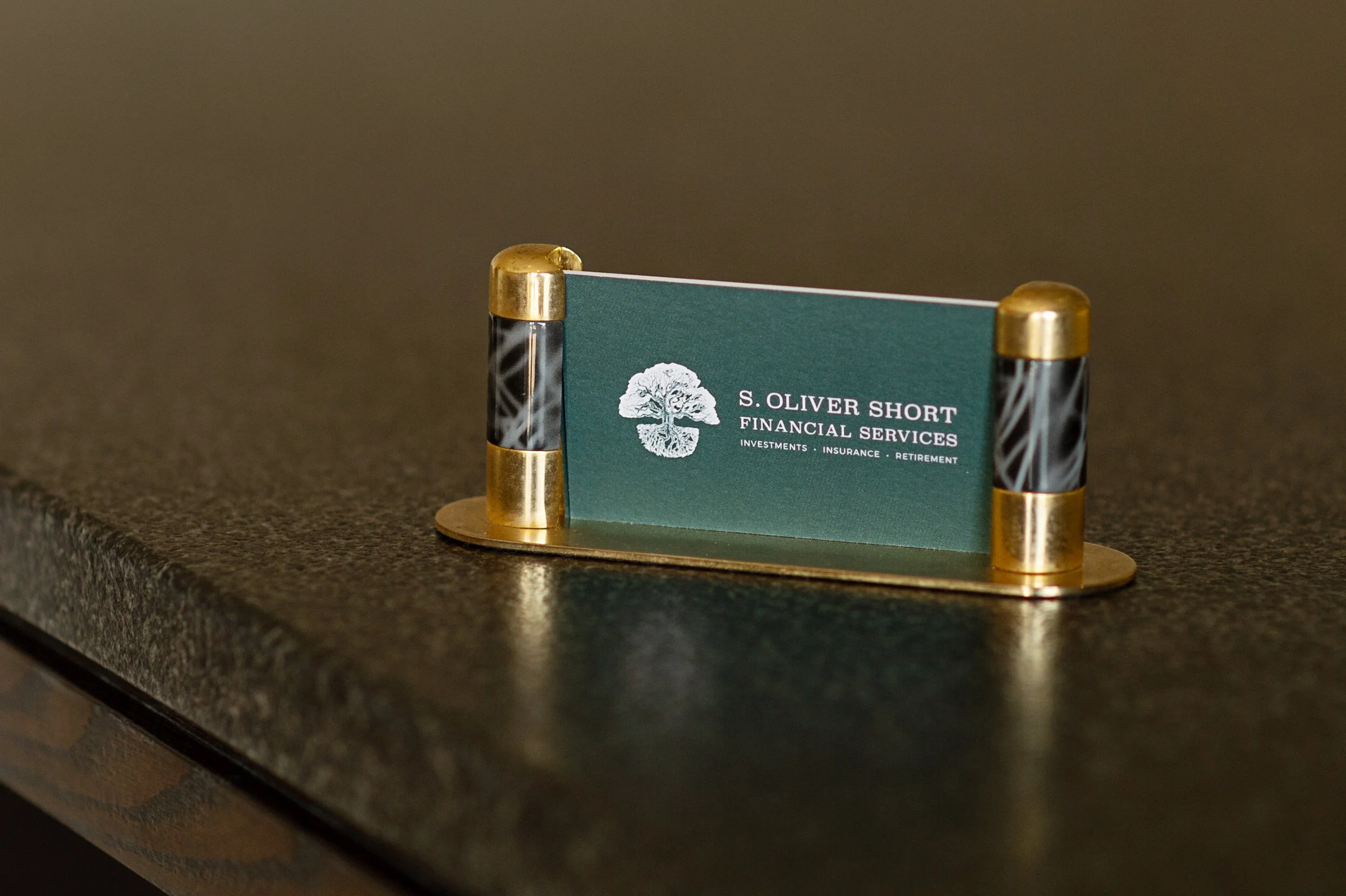 A business card for S. Oliver Short Financial Services, featuring a white tree logo on a green background, positioned on a small metal stand on a dark textured surface.
