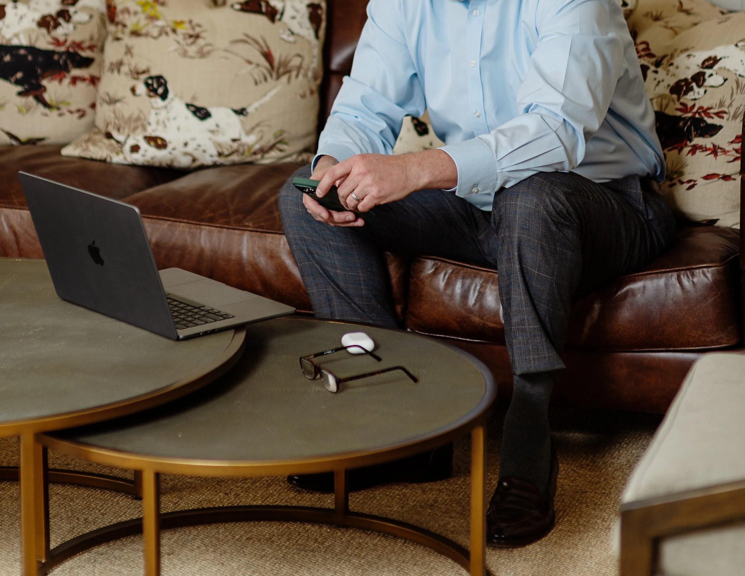 A person dressed in a light blue shirt and dark patterned pants, sitting on a brown leather couch, holding a smartphone. There are patterned pillows behind him, a laptop and eyeglasses on a round wooden table in front of him, and a pair of glasses on the table.