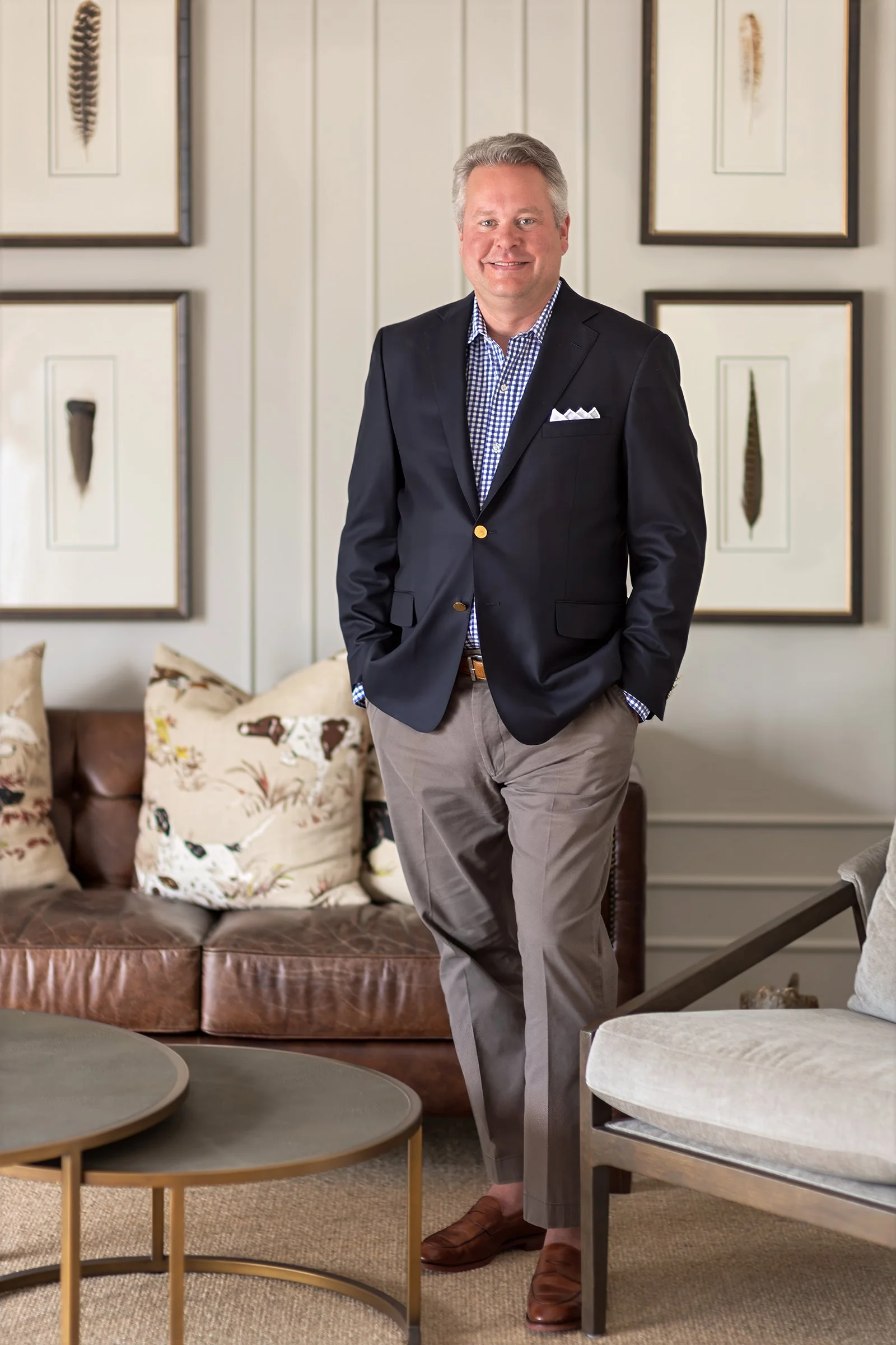 A man in a dark blazer, checked shirt, beige pants, and brown shoes standing in a modern living room with art on the wall, a leather sofa with floral pillows, and round nesting Turkish coffee tables.