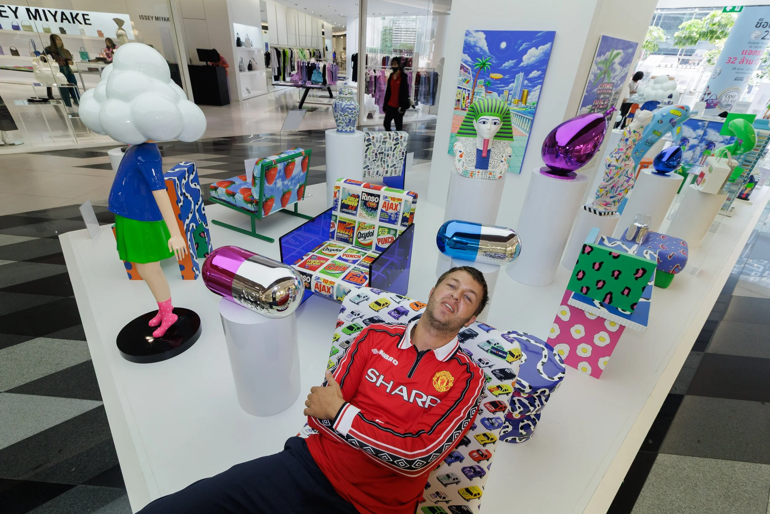 A man wearing a red Manchester United jersey and another person are posing at a display of colorful art sculptures and furniture inside a shopping mall.