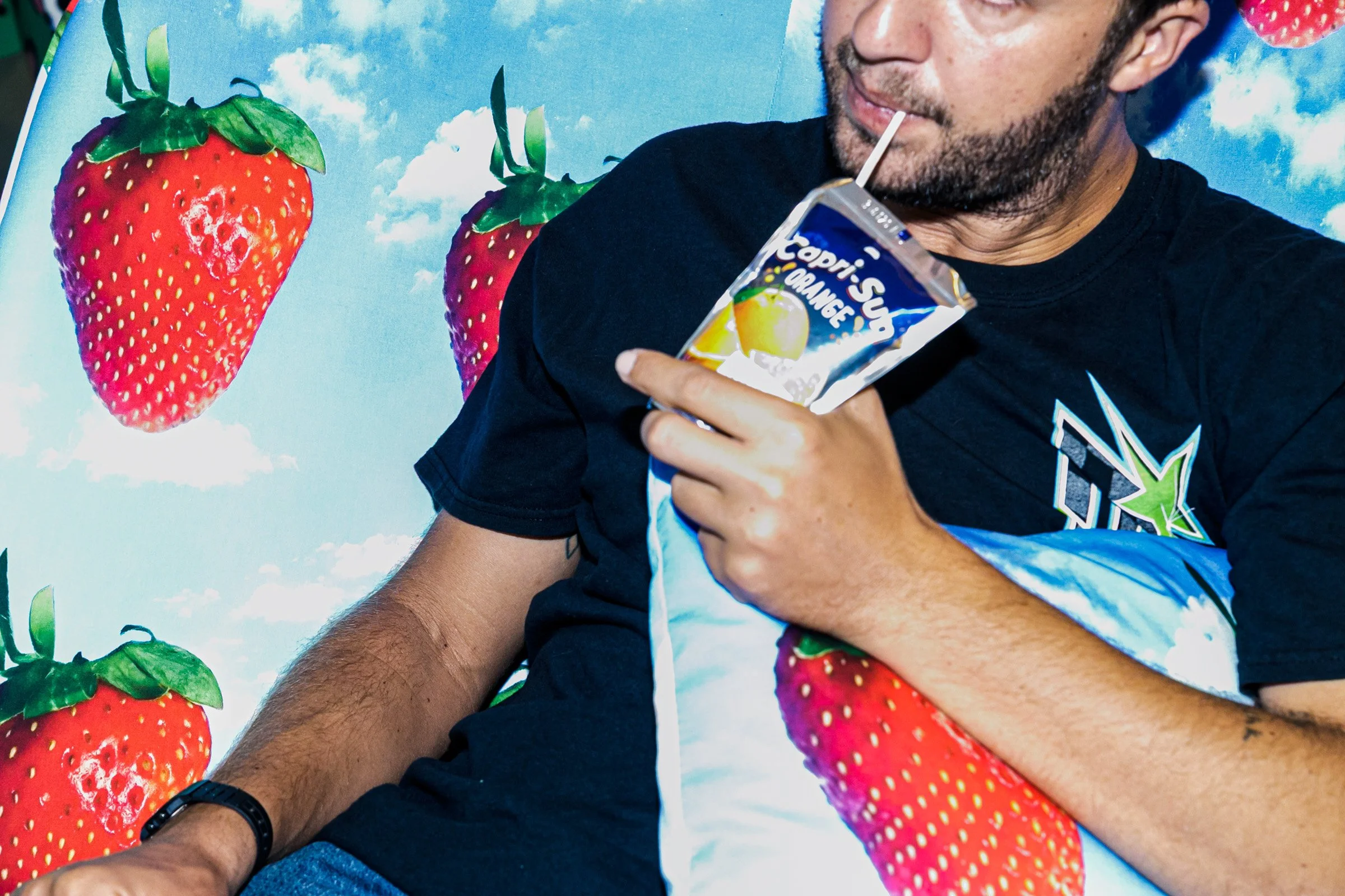 A man with a beard is sucking on a straw from a small food or drink package, with a background featuring large strawberries and a sky with clouds.