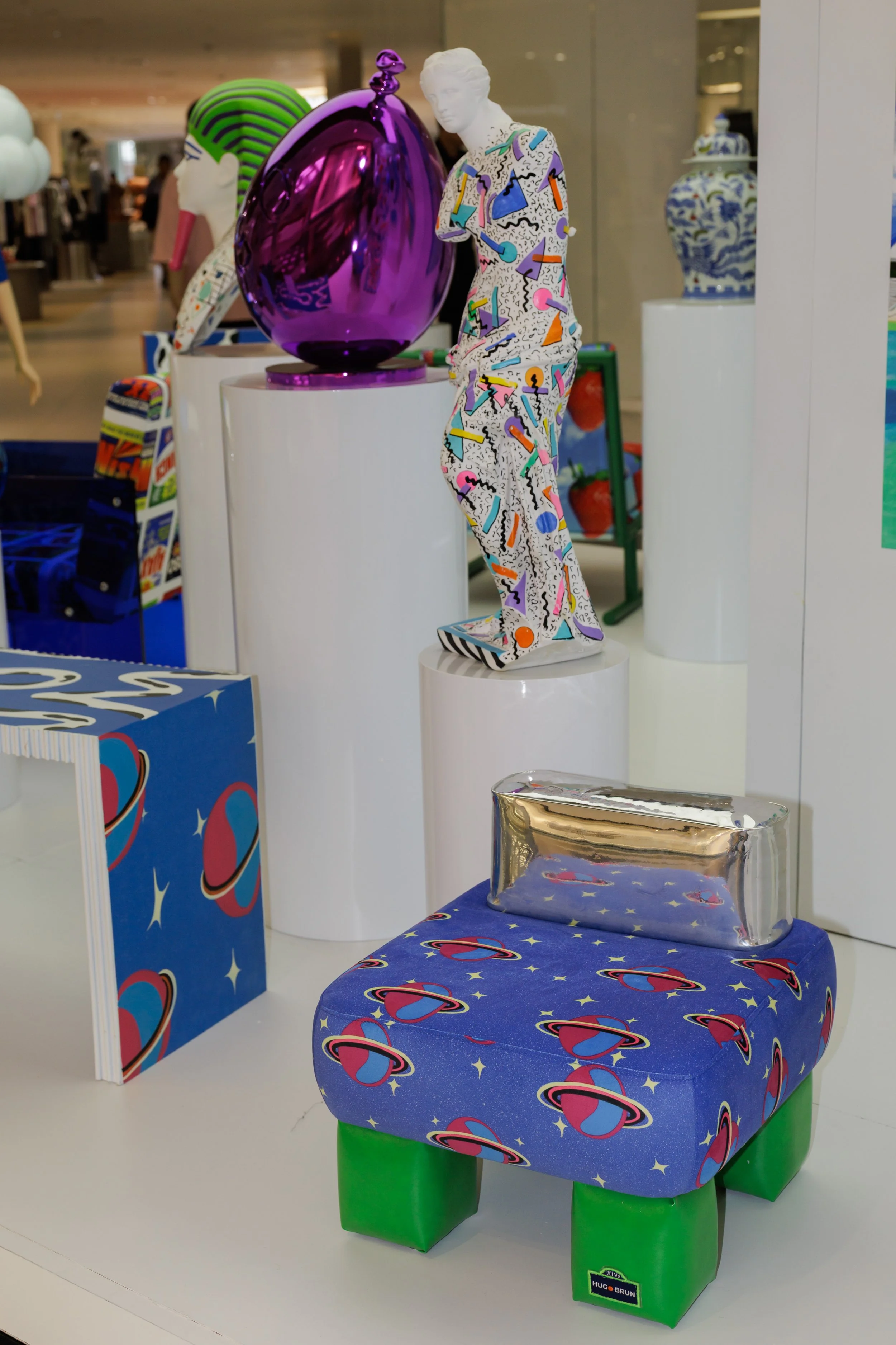 Colorful display of modern art sculptures in a shopping mall. Includes a white mannequin with a vibrant pattern, a large purple egg-shaped sculpture, and other artistic pieces with bright and playful designs.