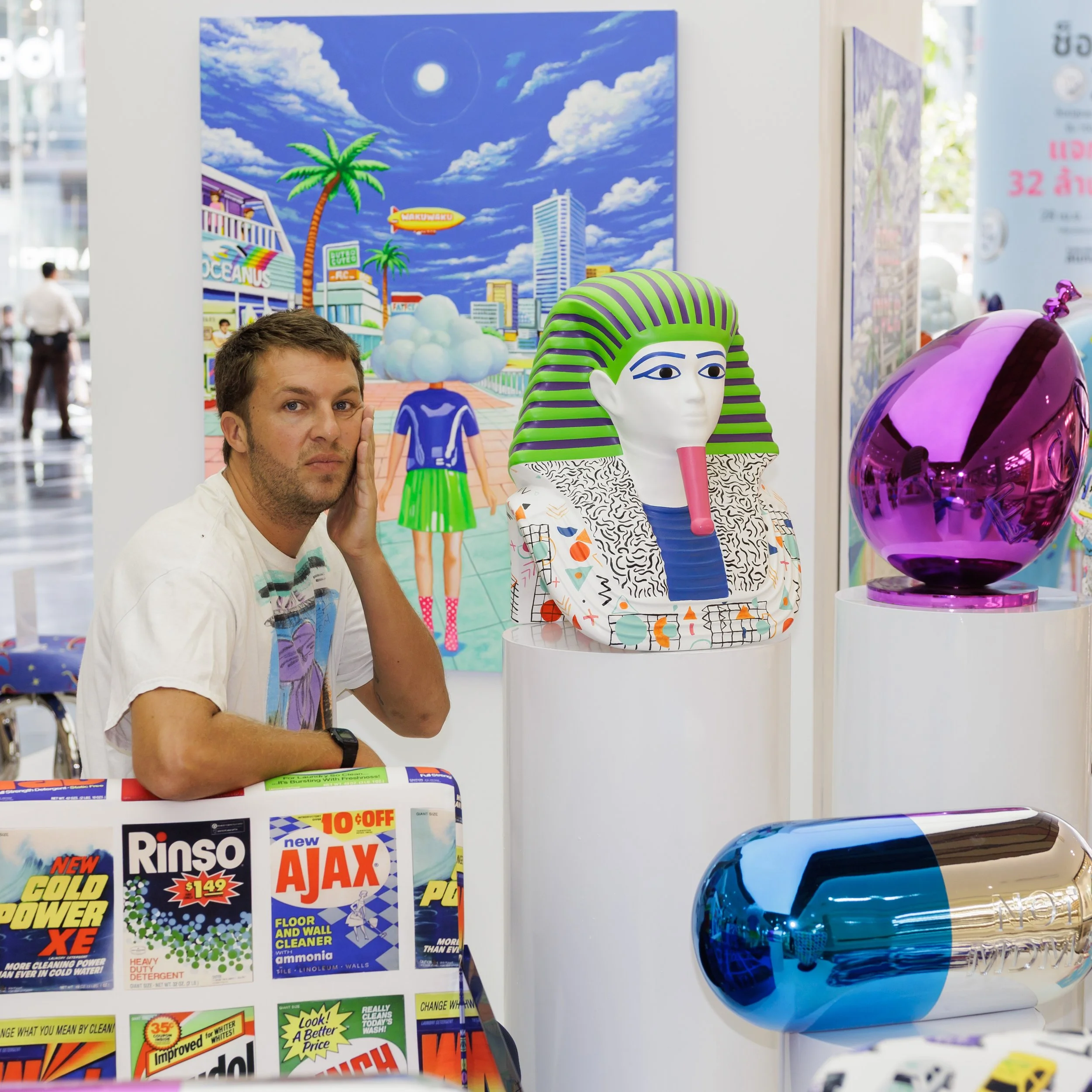 Close-up of a man with short brown hair and a beard holding his face near a display of colorful cleaning supplies, with a large blue and purple glass sculpture and vibrant posters in the background at an indoor store or exhibition.