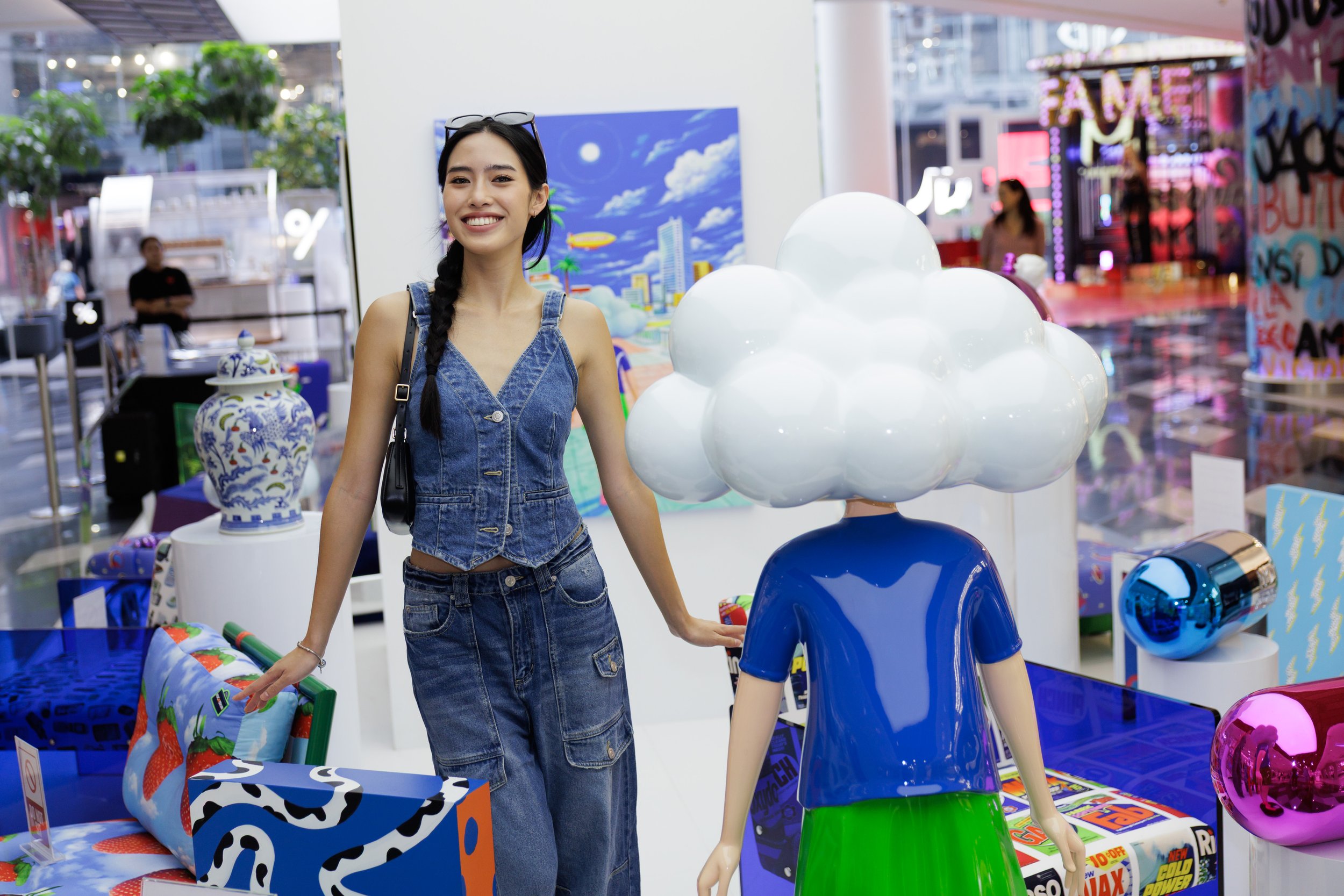 A woman smiling at a store display shelf with colorful items, standing next to a child-sized mannequin with a cloud-shaped head and a blue outfit, in a shopping mall.