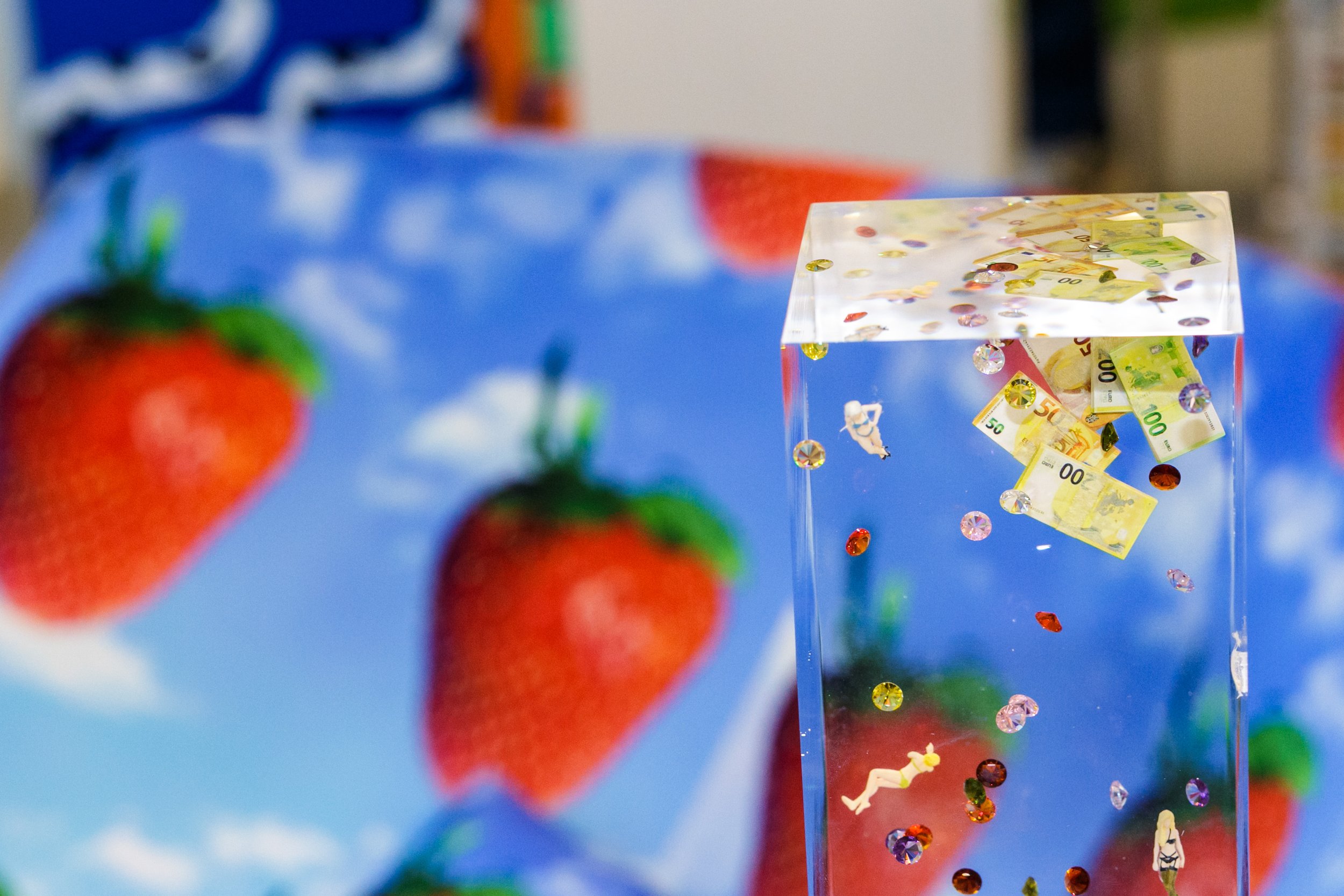 A transparent cube filled with inflatable Canadian dollar bills and colorful rhinestones, with blurred red strawberries in front and a blue background.