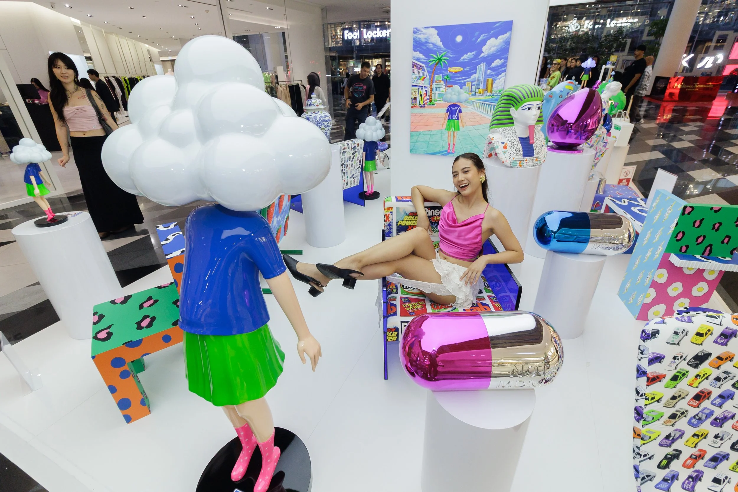 Two young women in a shopping mall display area with colorful, contemporary art and sculptures. One woman is sitting on a bench, smiling and wearing a pink top and white skirt, while the other woman, partially visible, stands beside a stylized manneq