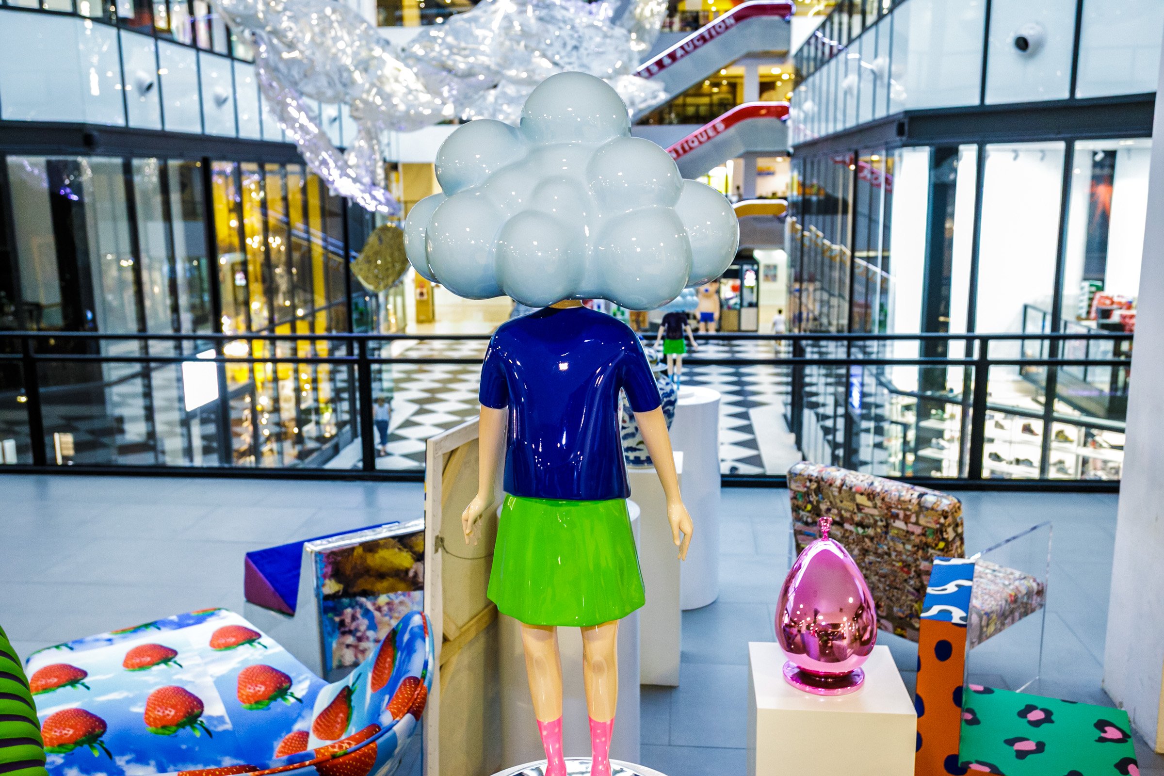 A mannequin with a cloud-shaped head made of white round bubbles, wearing a blue top and a green skirt, stands indoors in a shopping mall. Surrounding it are colorful wrapped gifts and decorative items, with glass railings, escalators, and store windows visible in the background.