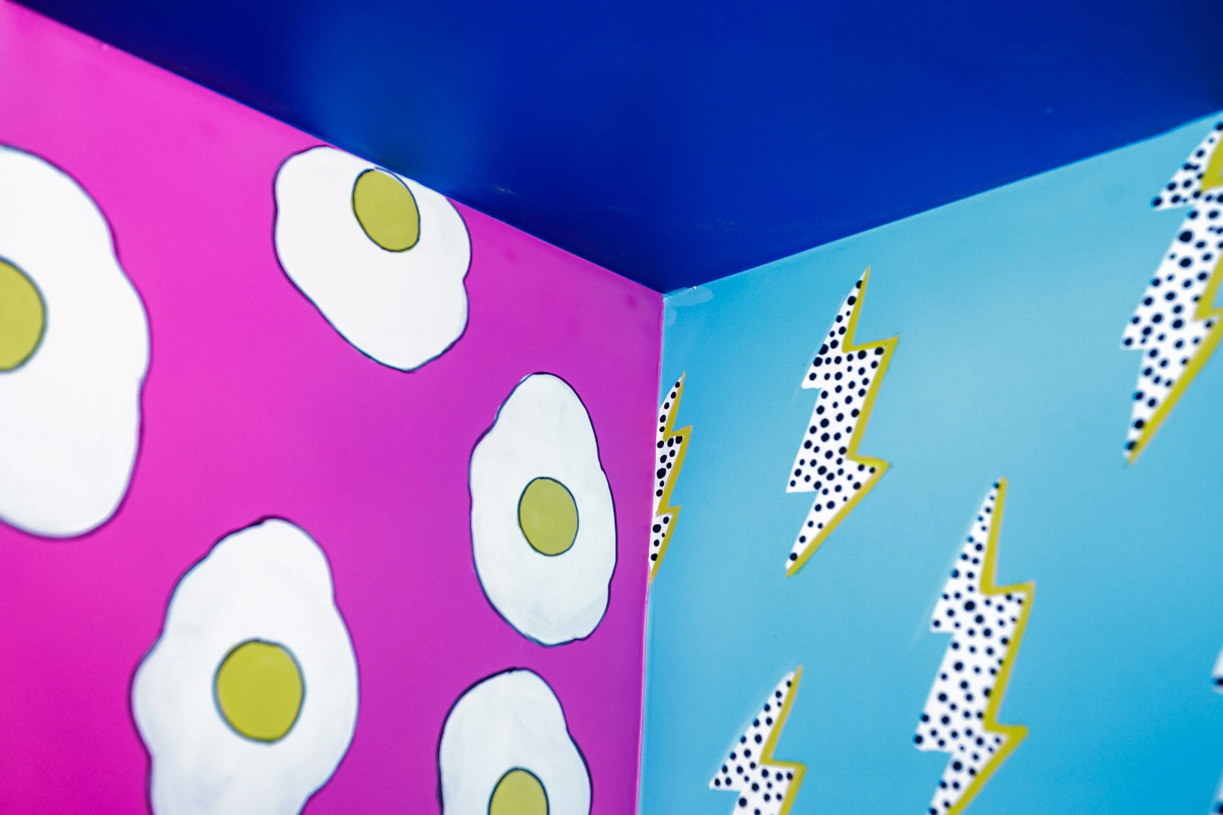 Close-up of a colorful box with pop art style patterns including white eggs with yellow yolks on a pink background and black and white polka dots with lightning bolt designs on a blue background.