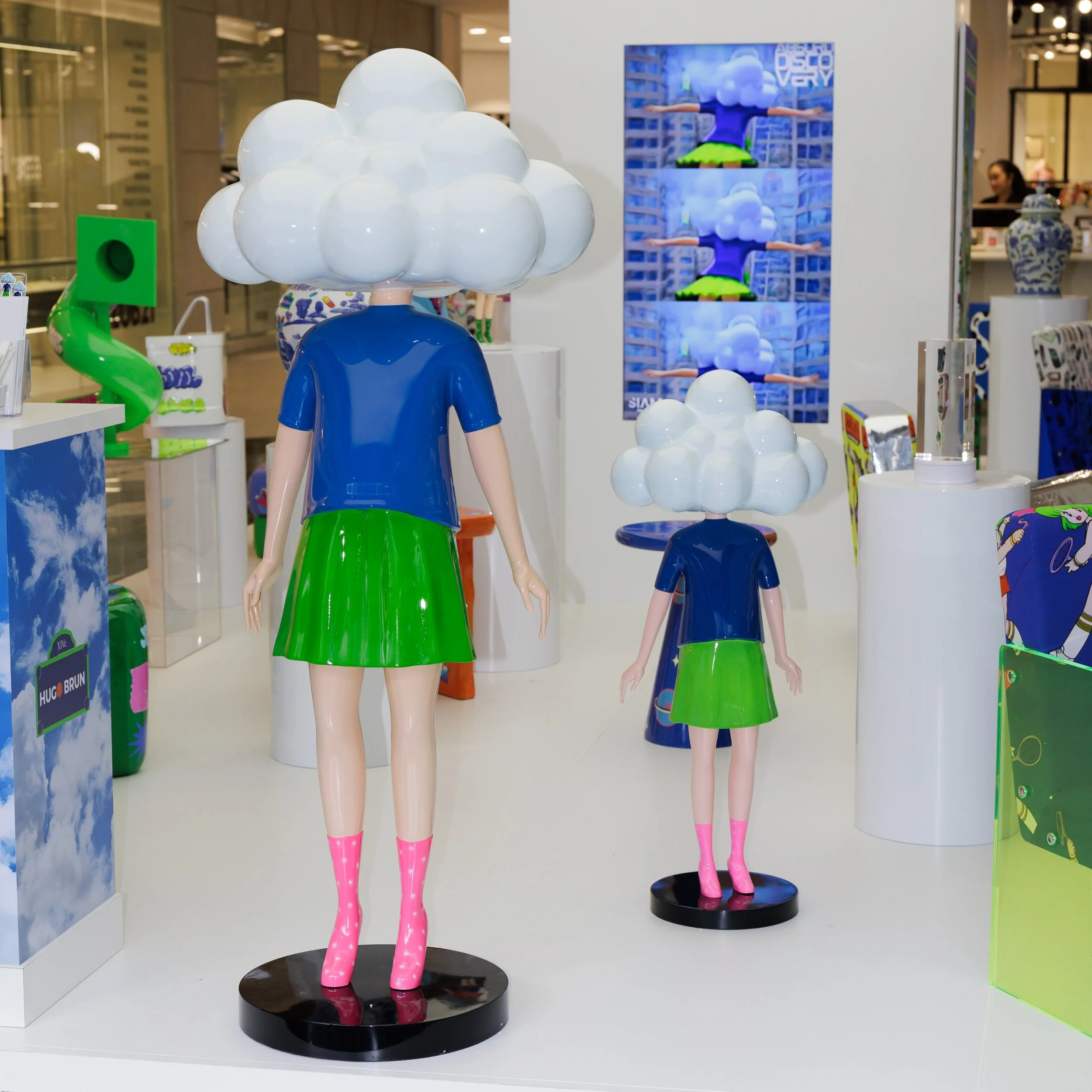 Mannequin with a large cloud-shaped head, wearing a blue shirt and a green skirt, pink rain boots, displayed in a retail store exhibit.