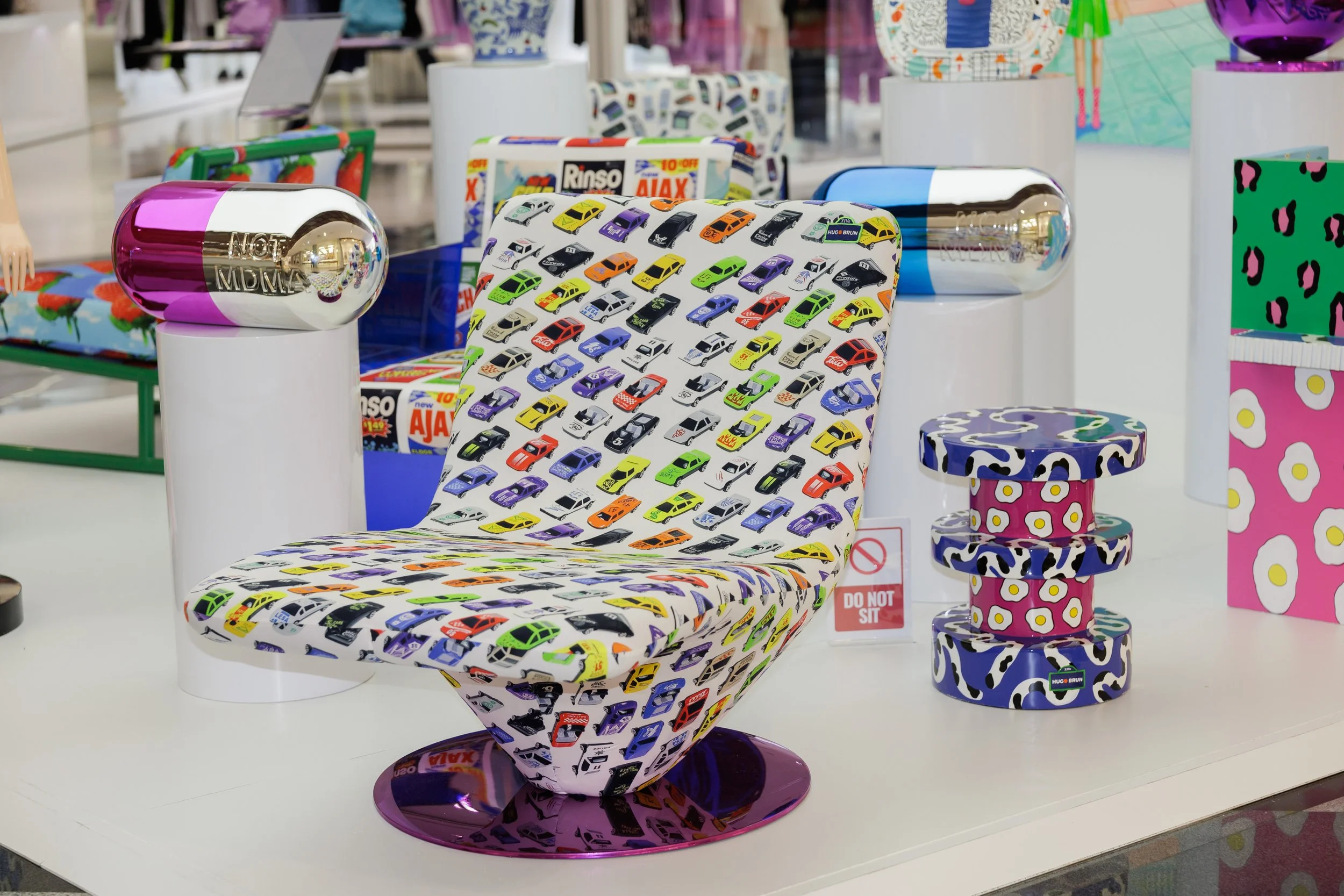 Display of colorful furniture and decor items in a store, including a chair with car print upholstery, a stool with a pink and white pattern, and various patterned boxes and decorative objects.