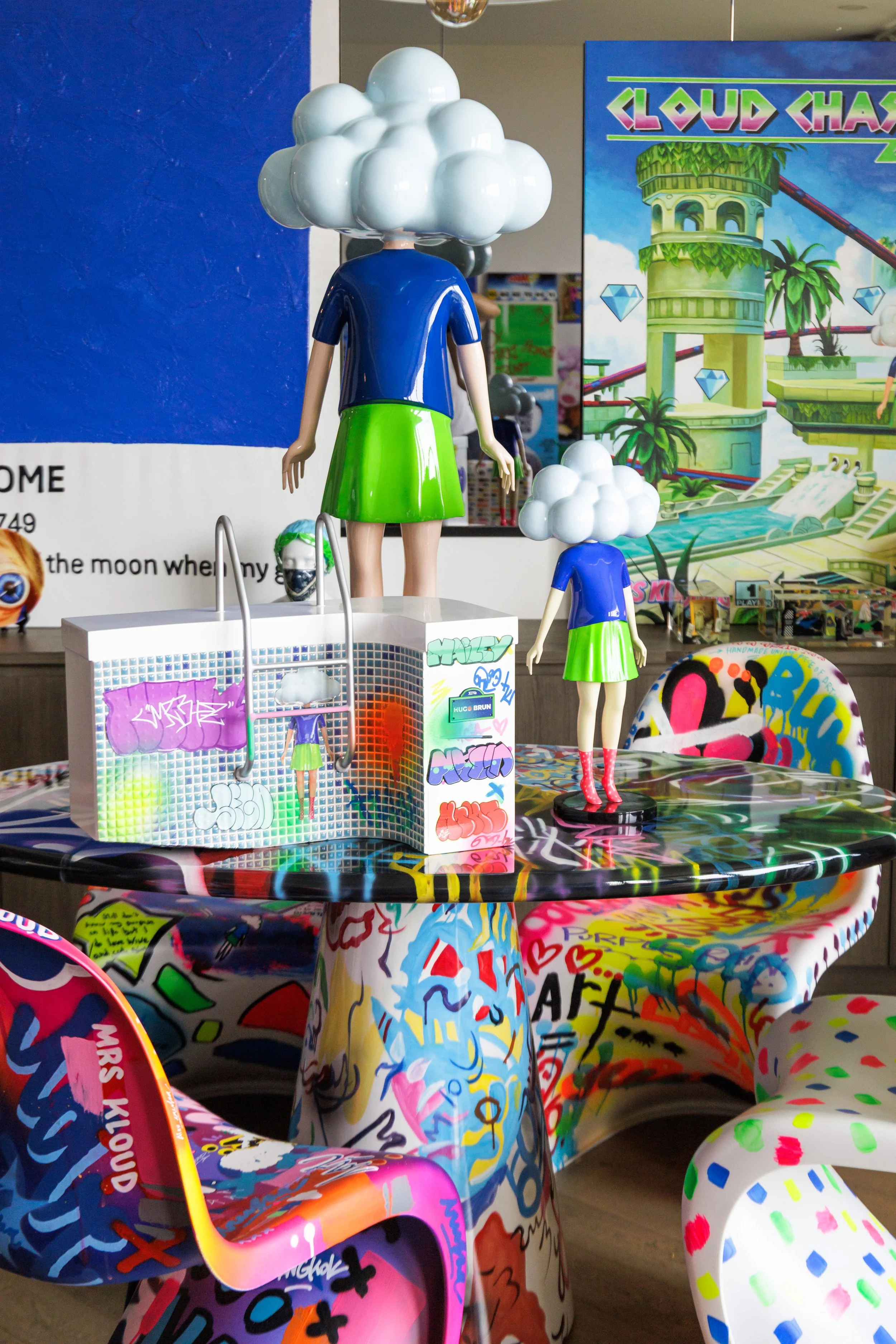 Colorful art installation with mannequin figures that have cloud-shaped heads, wearing blue shirts and green skirts, displayed on a decorated table with graffiti-style patterns.